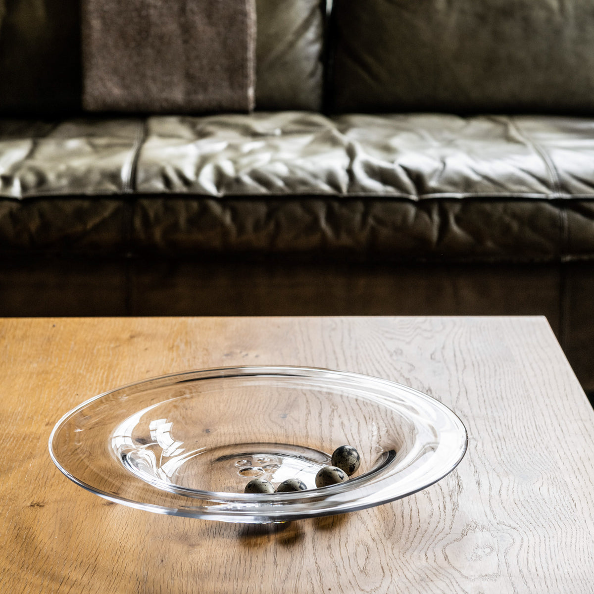 Clear glass serving platter with olives on wooden table in front of dark leather sofa.