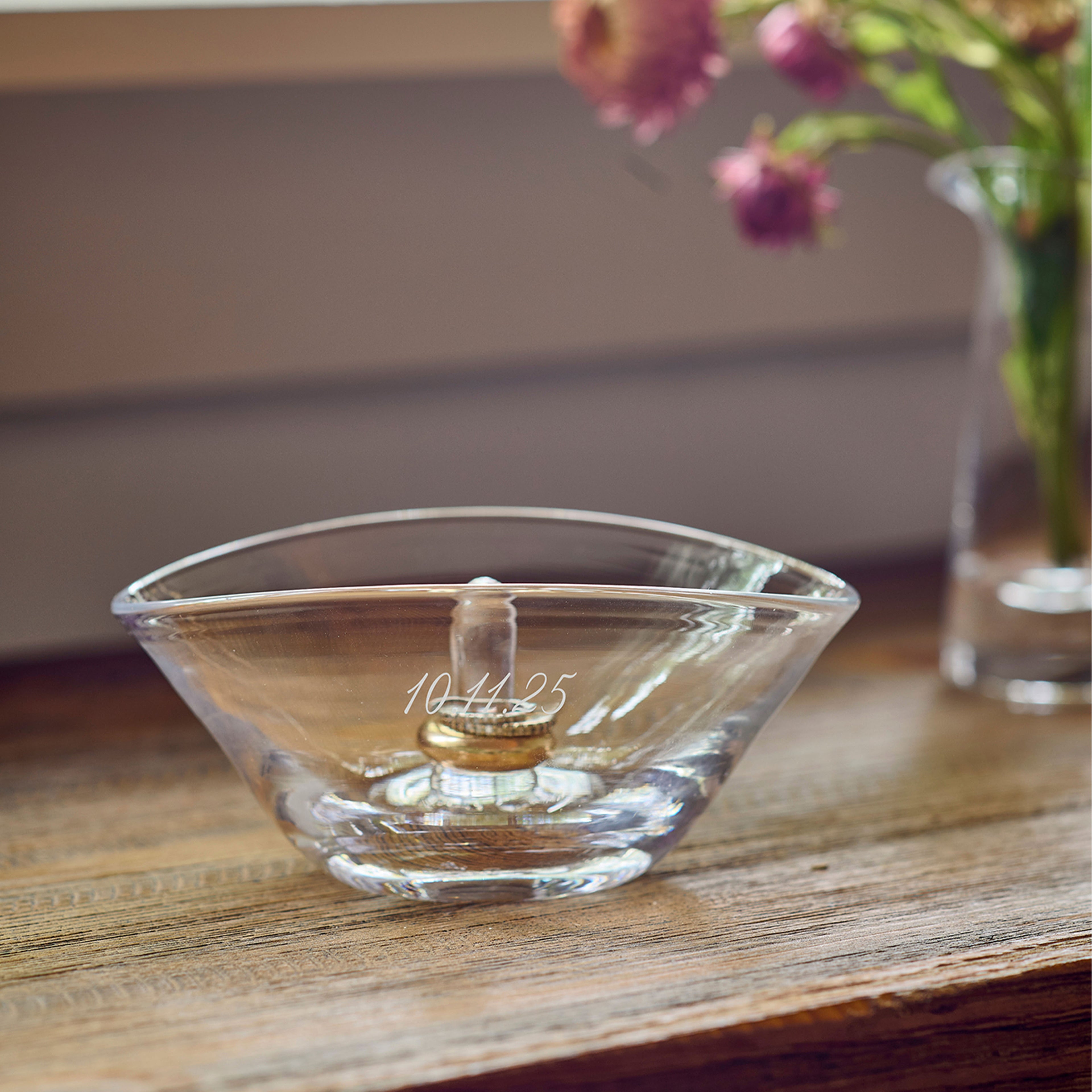 Clear glass bowl with wide oval opening and engraved text on front, holding a gold ring on interior post.