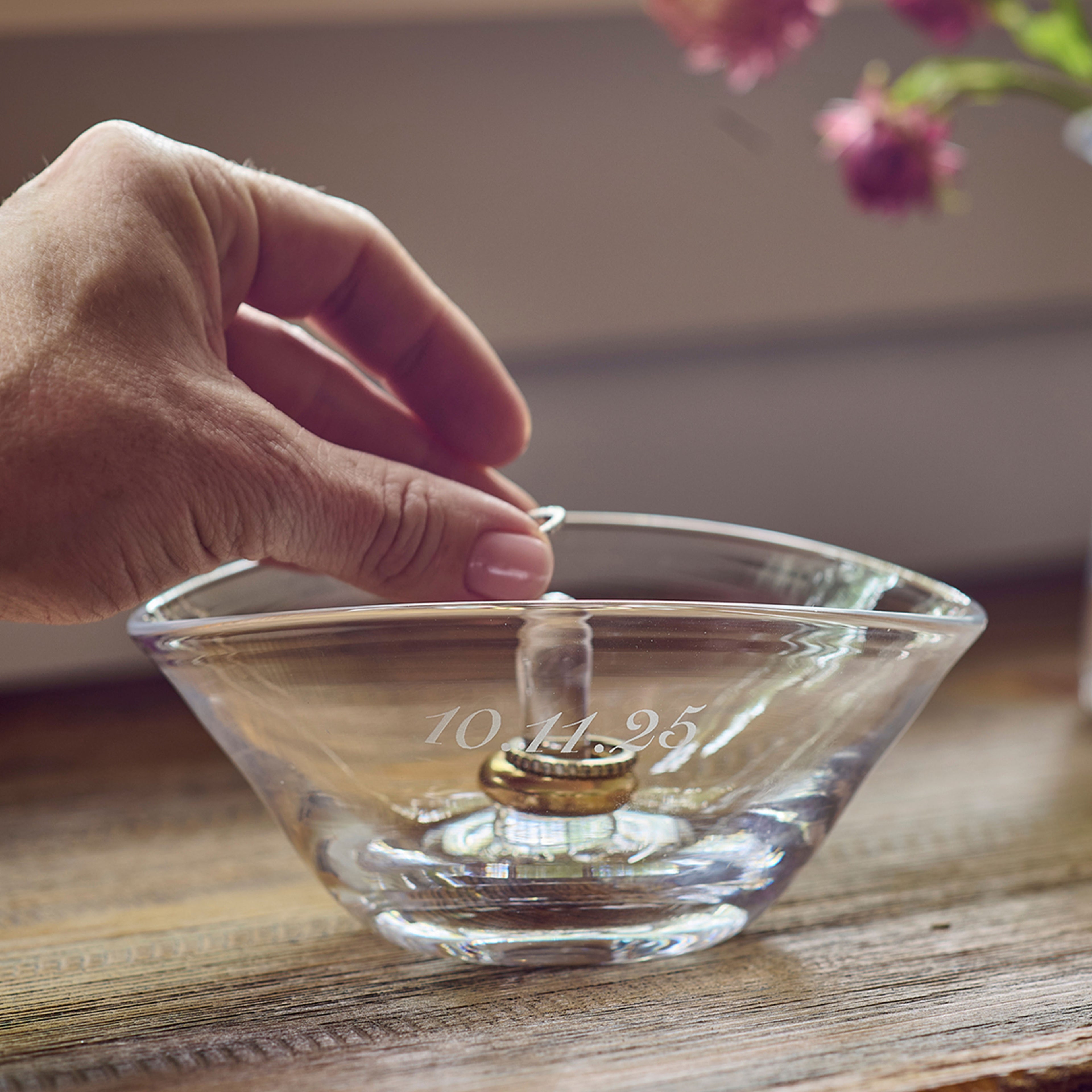Clear glass ring holder with engraved date, hand placing ring on central post.