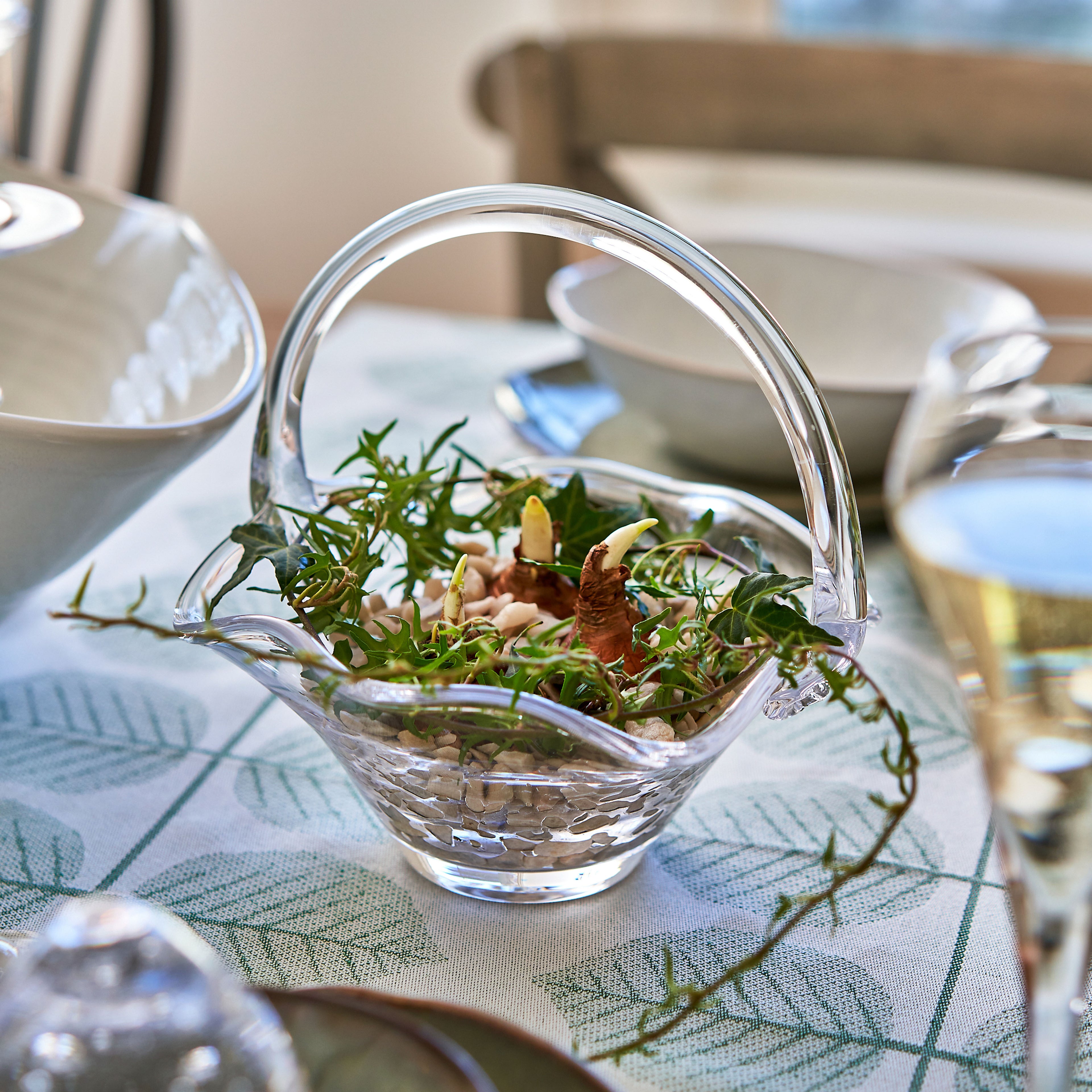 Clear glass basket with arched handle containing green foliage and small flowers on a patterned tablecloth.