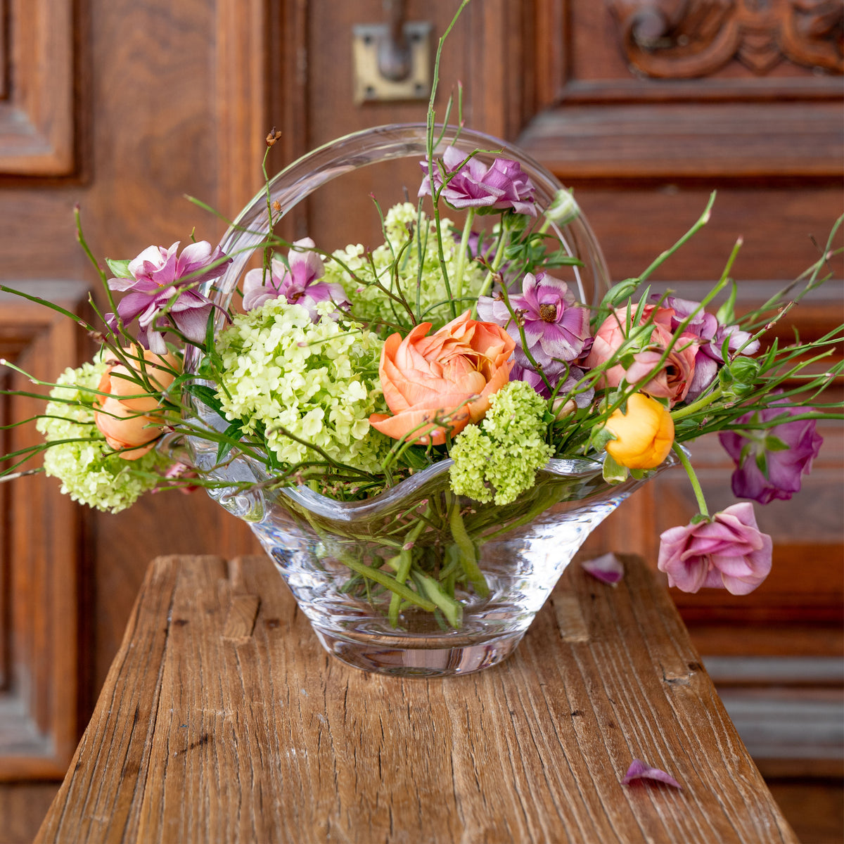 Clear glass basket-style vase filled with pink carnations, white hydrangea, orange roses, and green foliage on wooden surface