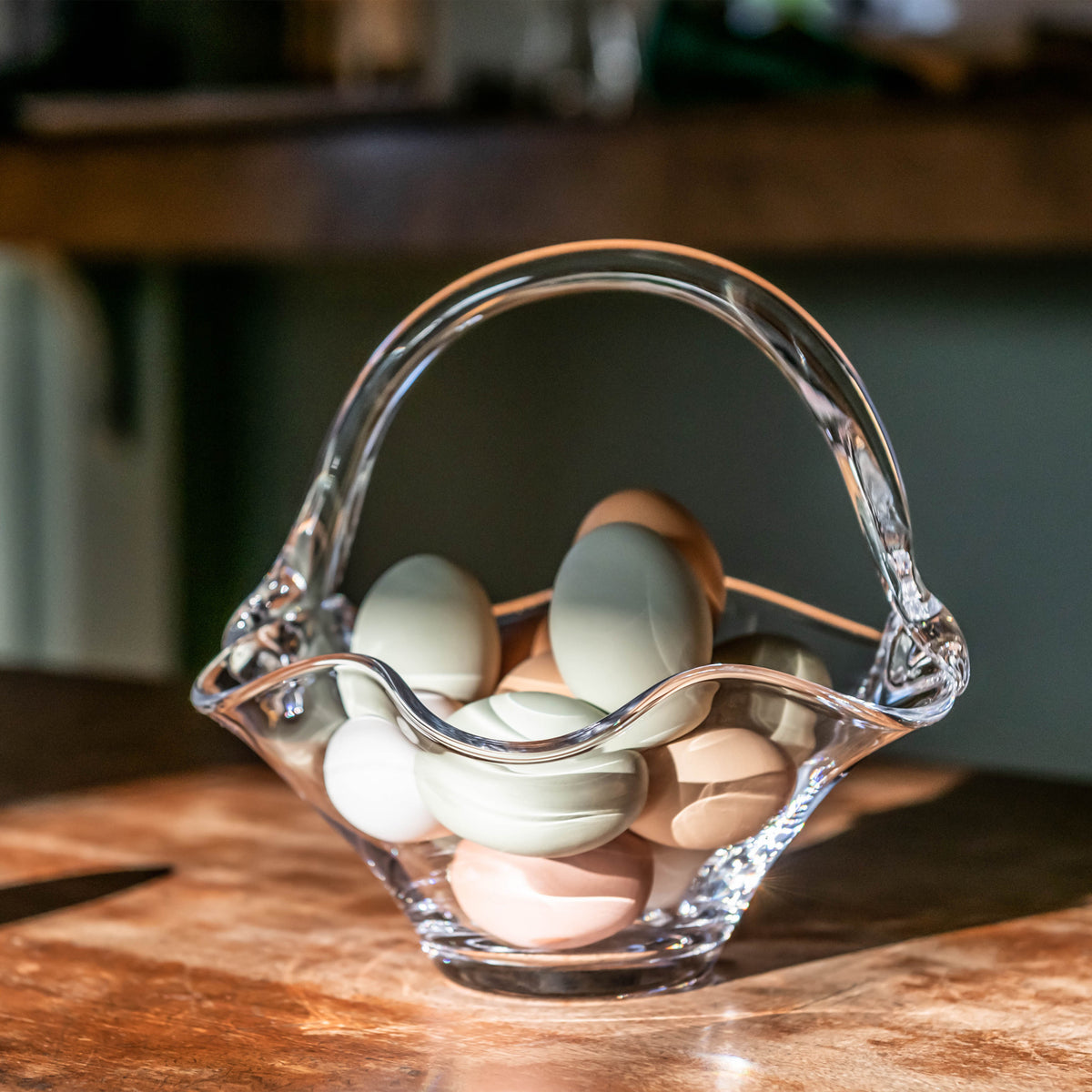 Clear glass basket with wavy rim and handle holding white and brown eggs on wooden surface.
