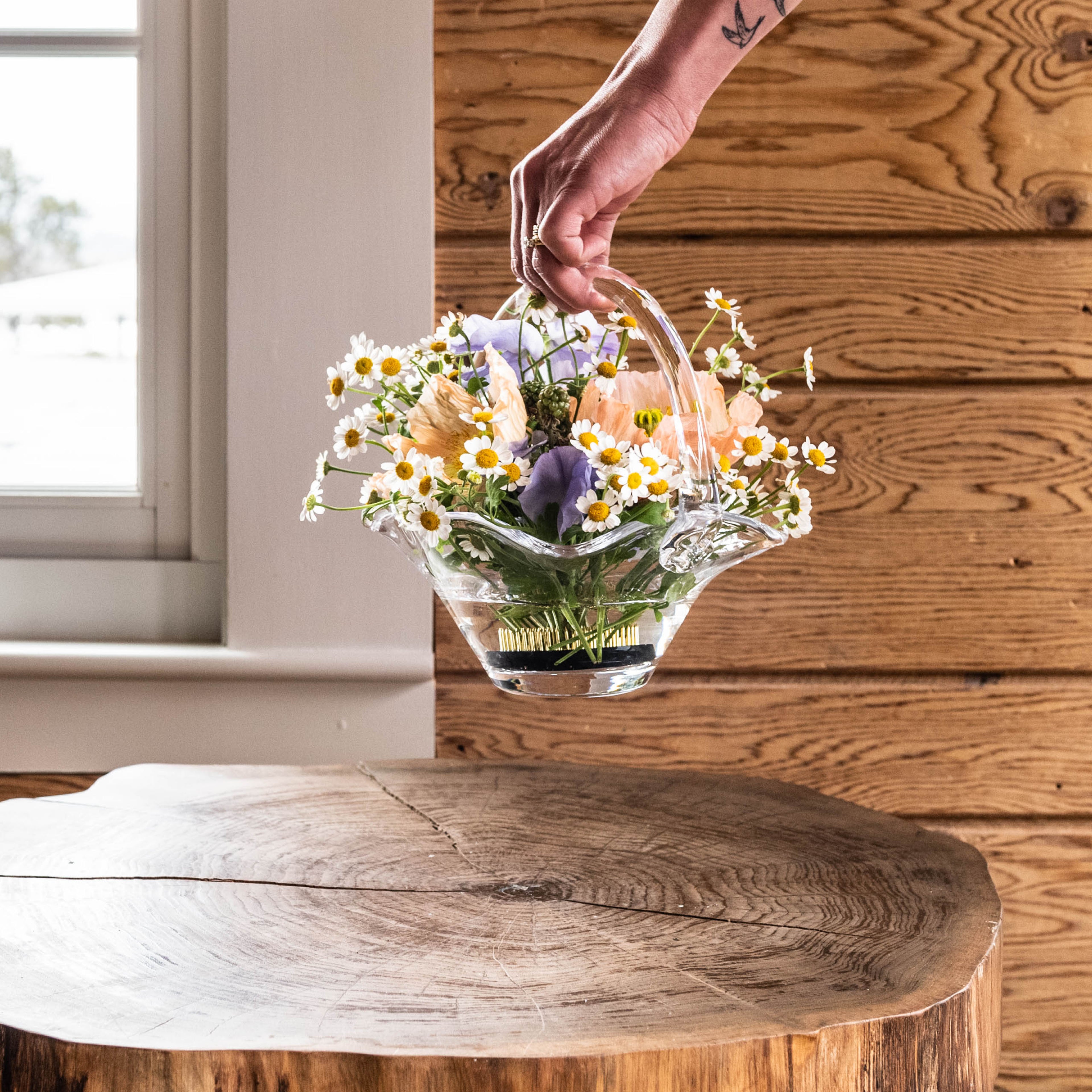 Clear glass basket with handles holding mixed daisies and purple flowers with green stems.