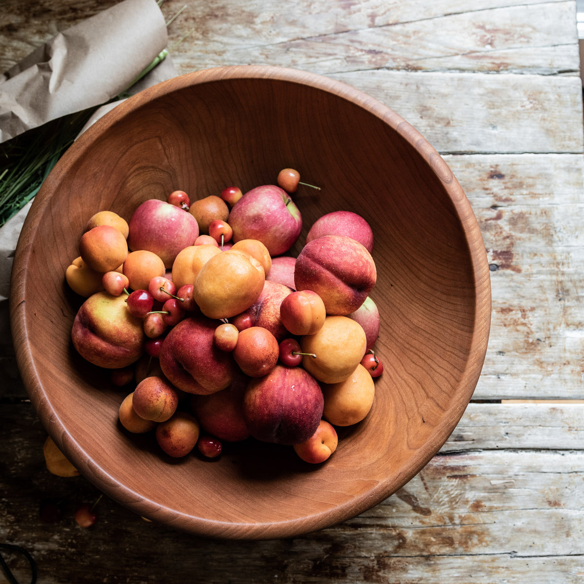 Cherry wood bowl with warm reddish-brown finish containing assorted fresh fruits on whitewashed wooden surface.
