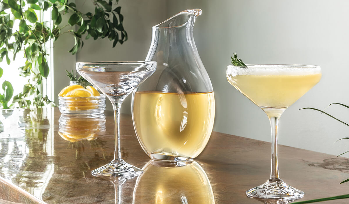 Clear glass carafe with pale yellow liquid, surrounded by matching cocktail glasses and fresh garnishes on wooden surface.