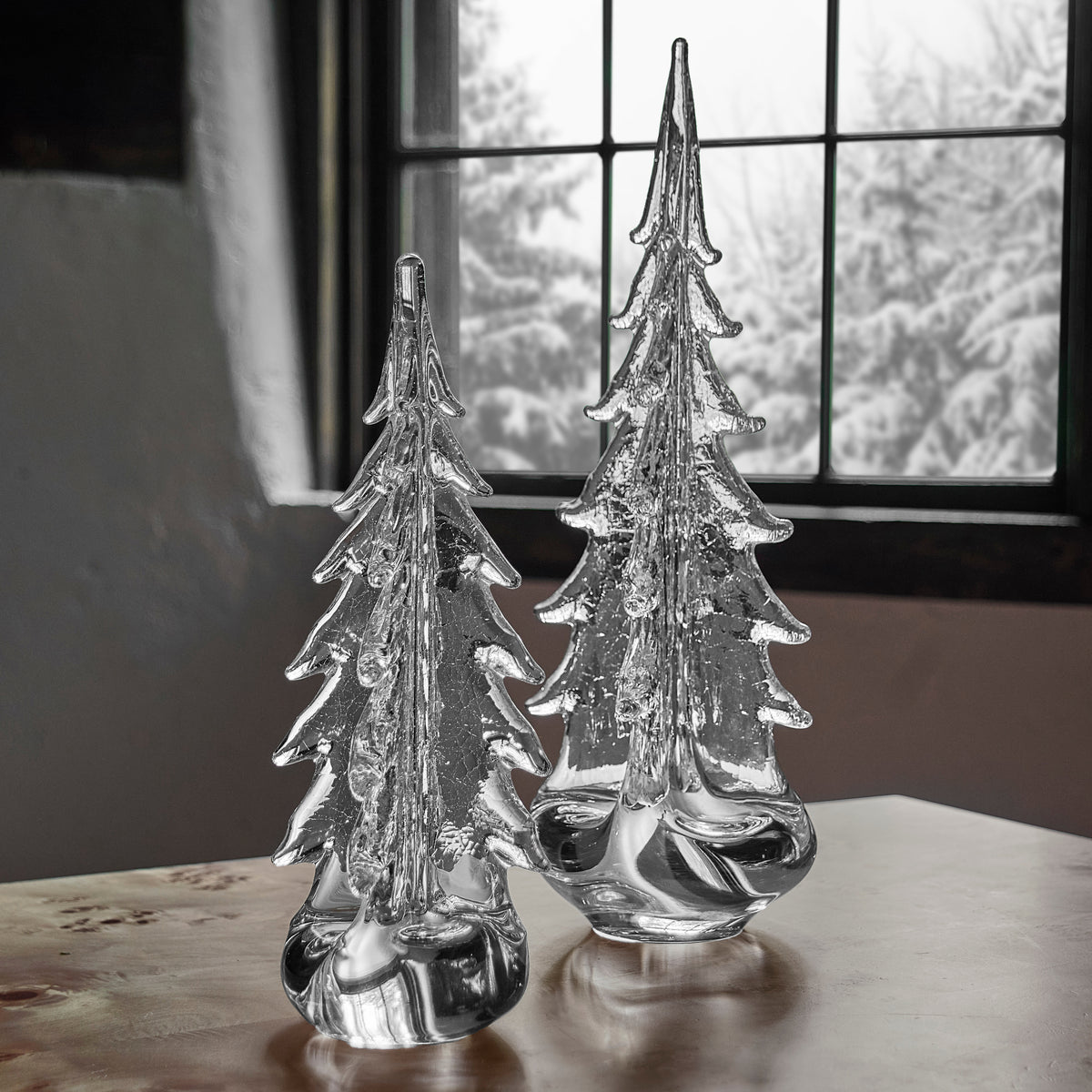 Two clear glass evergreen trees with textured crackle finish and pointed tops on round bases, displayed on a wooden surface.