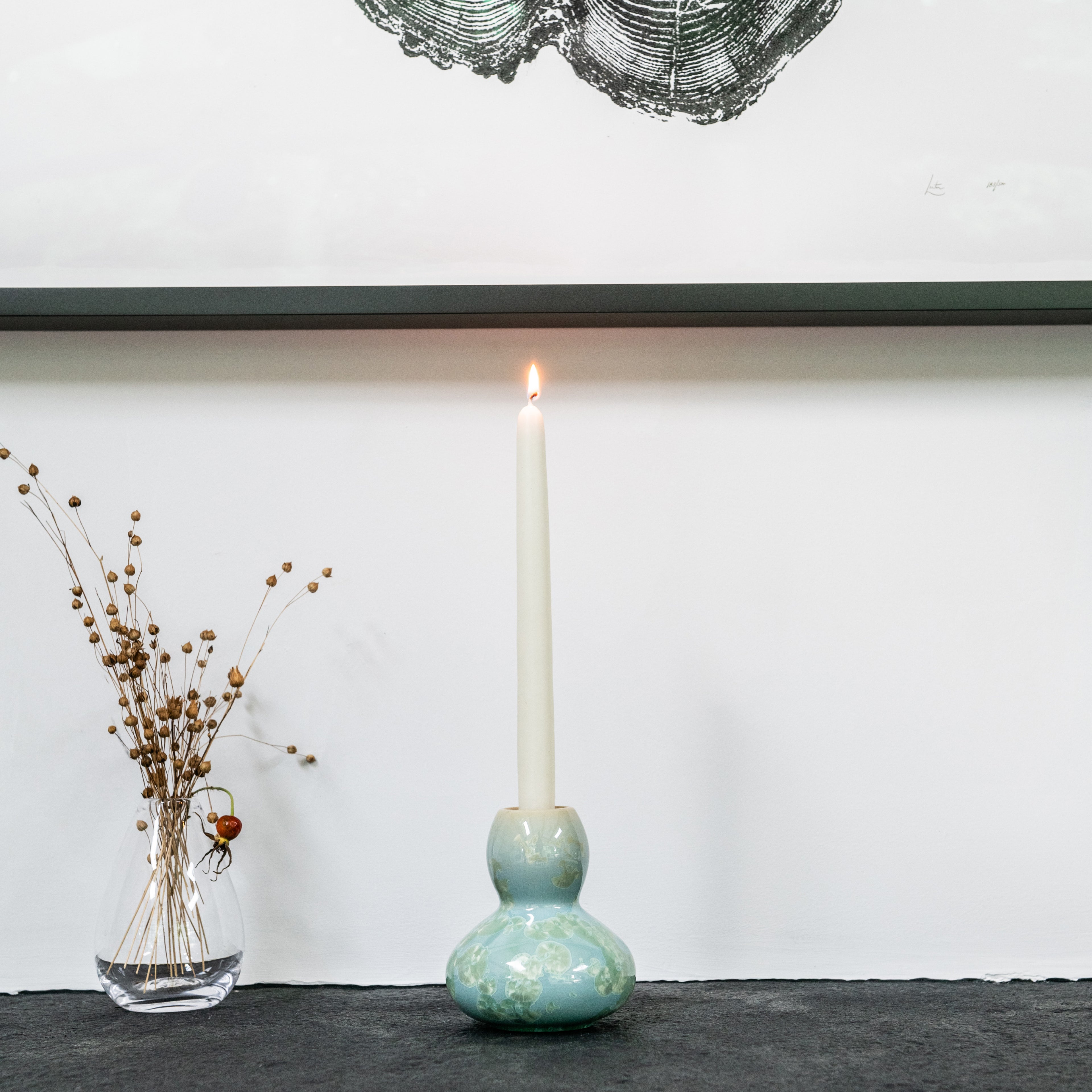 Ceramic candlestick with bulbous base in muted green glaze holding a lit cream taper candle.
