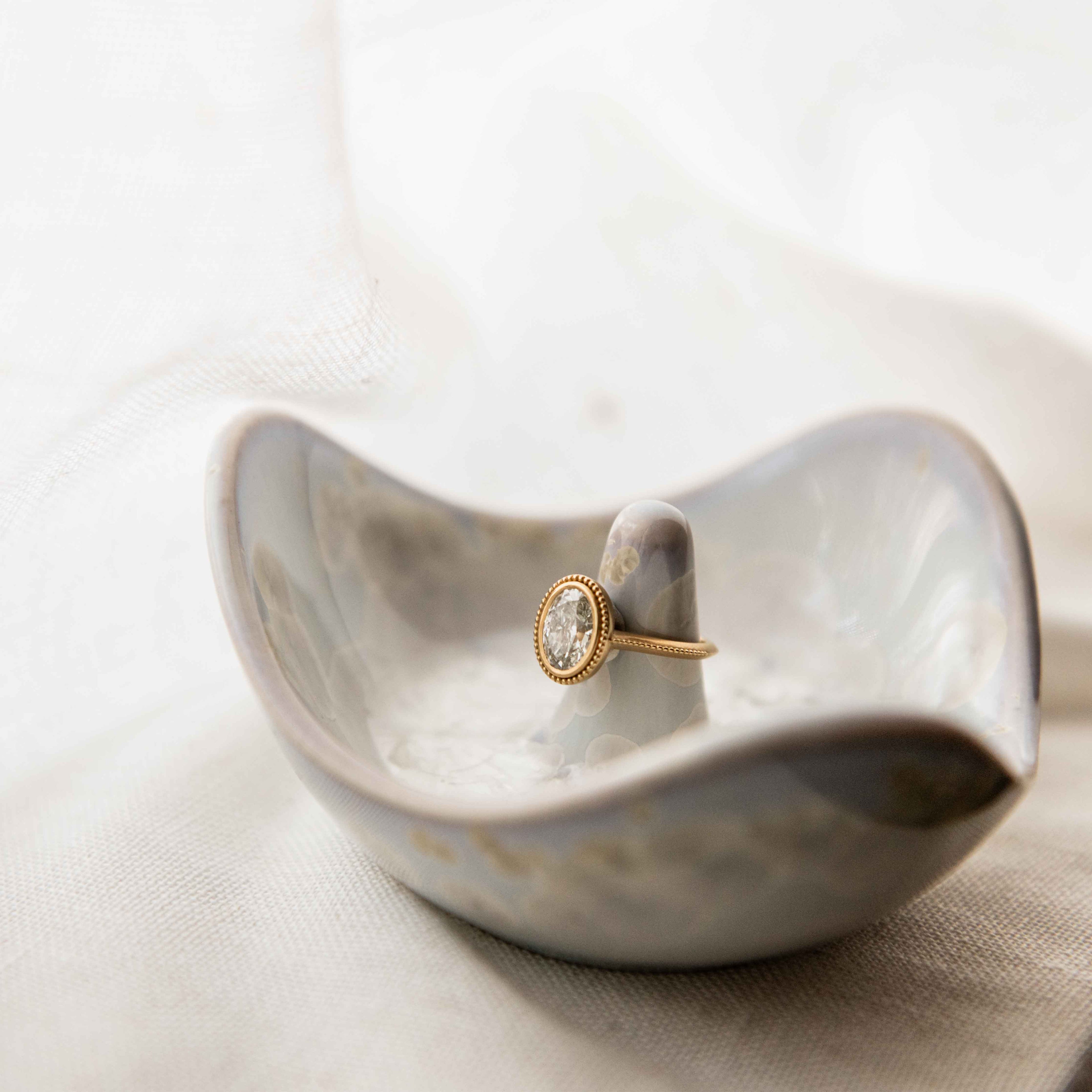 Gold ring with oval crystal stone displayed in white ceramic holder on neutral fabric surface.
