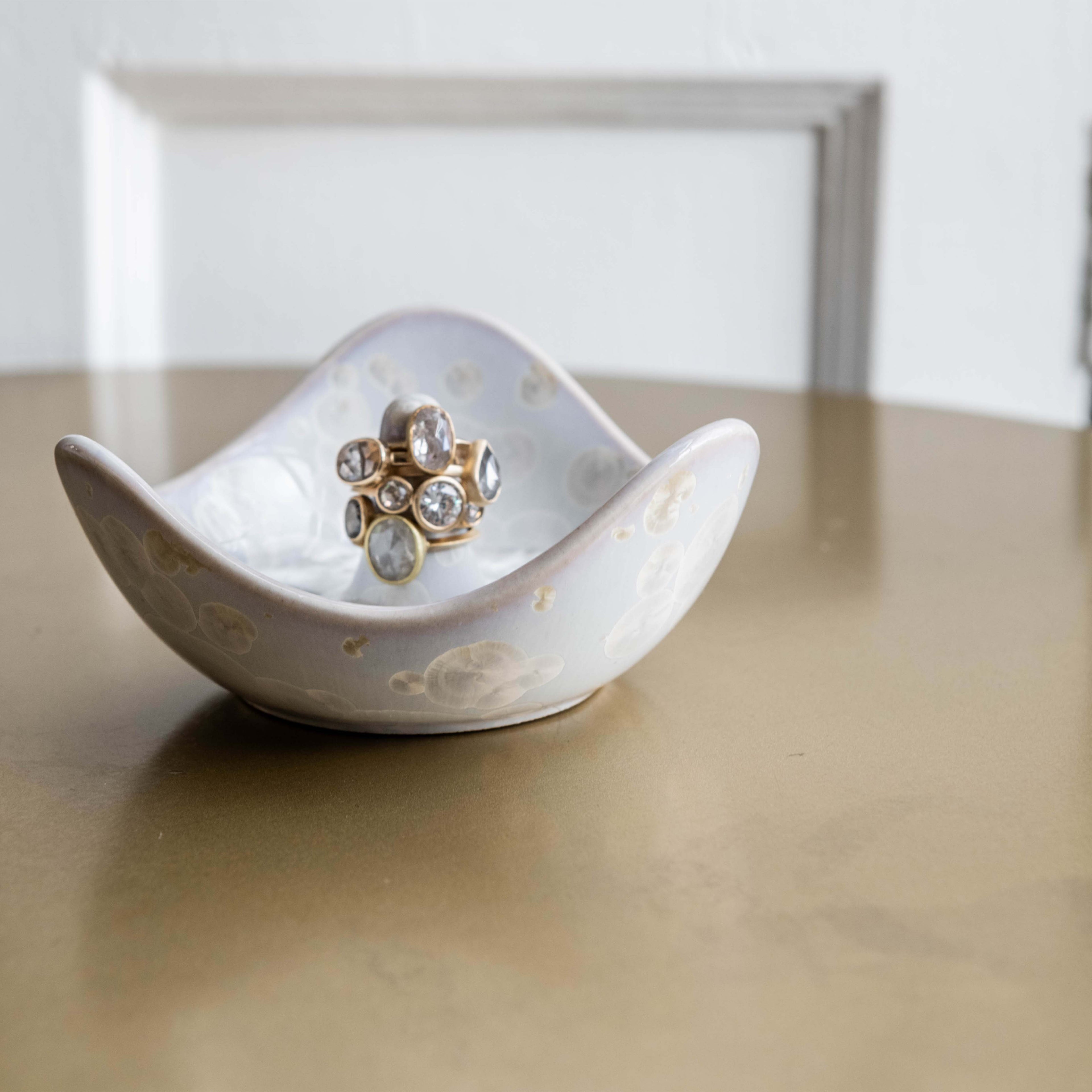 White crystalline ring holder with curved sides holding a gold-toned ring with clear stones, displayed with gift box in backg