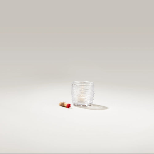 Clear glass tealight holder with textured surface and lit white candle inside, shown with red and gold match stick.