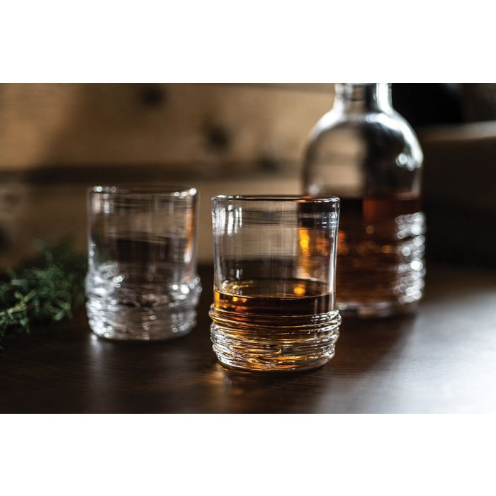 Two whiskey glasses with one containing amber liquid, empty glass beside it, and whiskey bottle in background with green herb