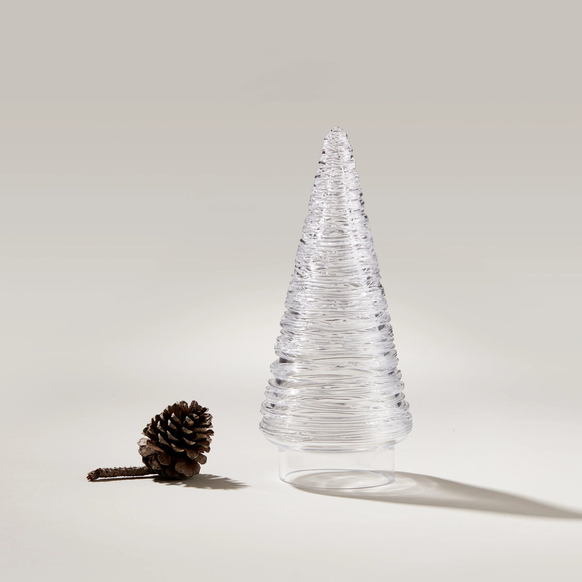 Clear glass cone-shaped tree with horizontal texture lines next to a brown pinecone on neutral background.