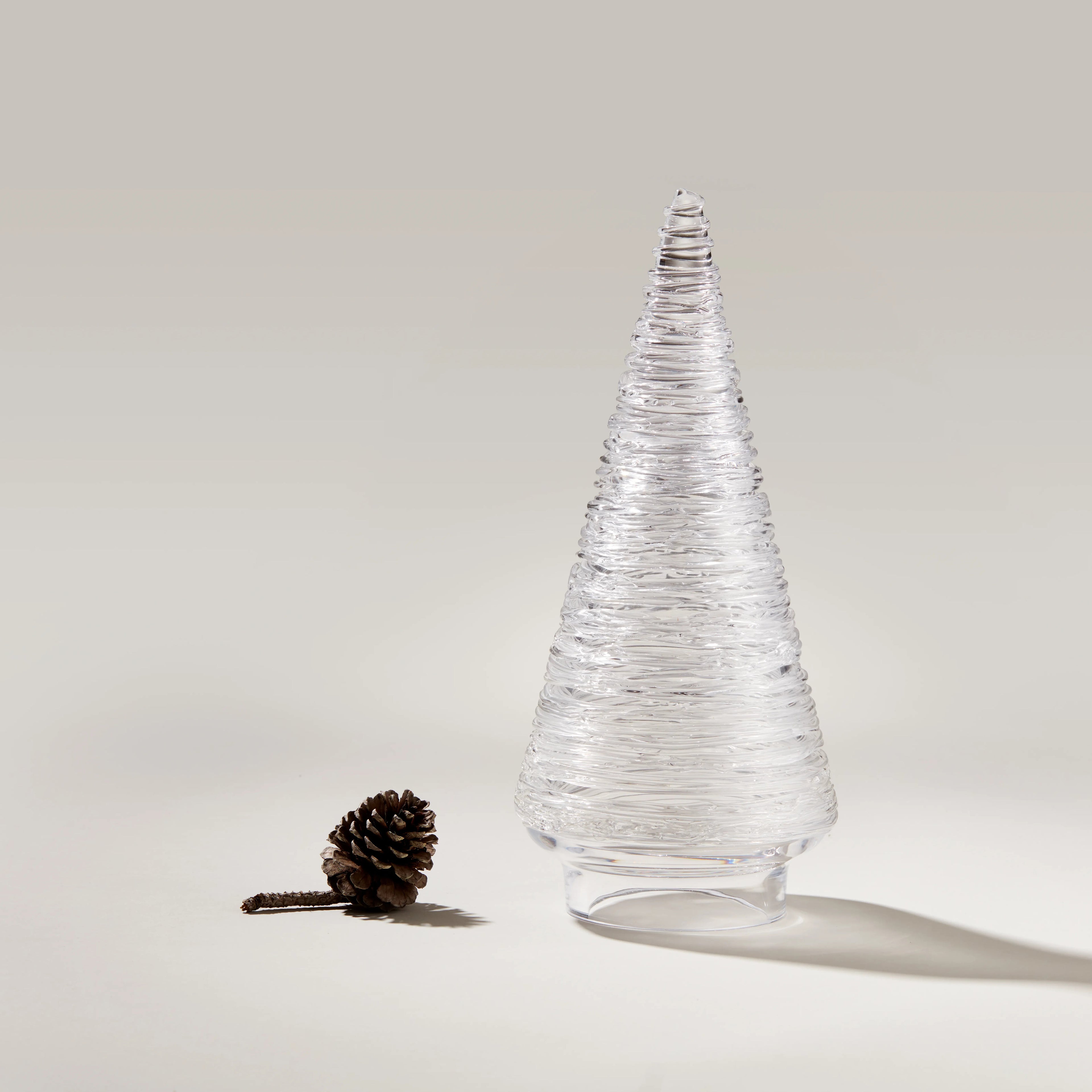 Clear glass cone-shaped decorative tree with textured surface and pinecone beside it on white background.