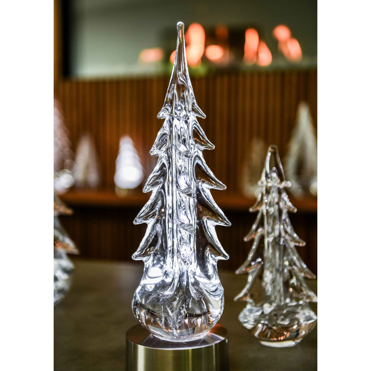 Clear glass evergreen tree with pointed top and tiered branches on metal base, blurred warm lights in background.