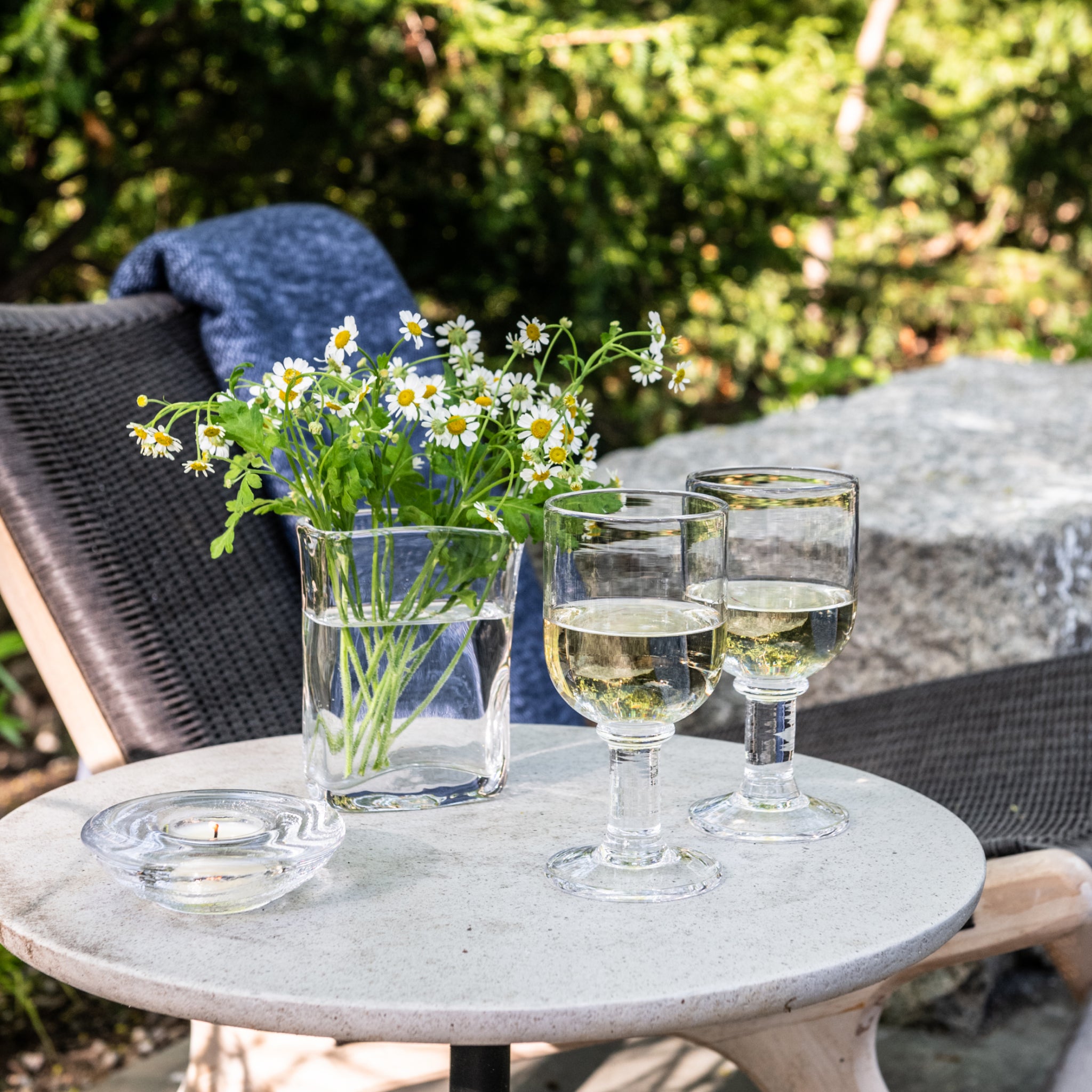 Two wine glasses filled with white wine on outdoor table with daisies in vase.