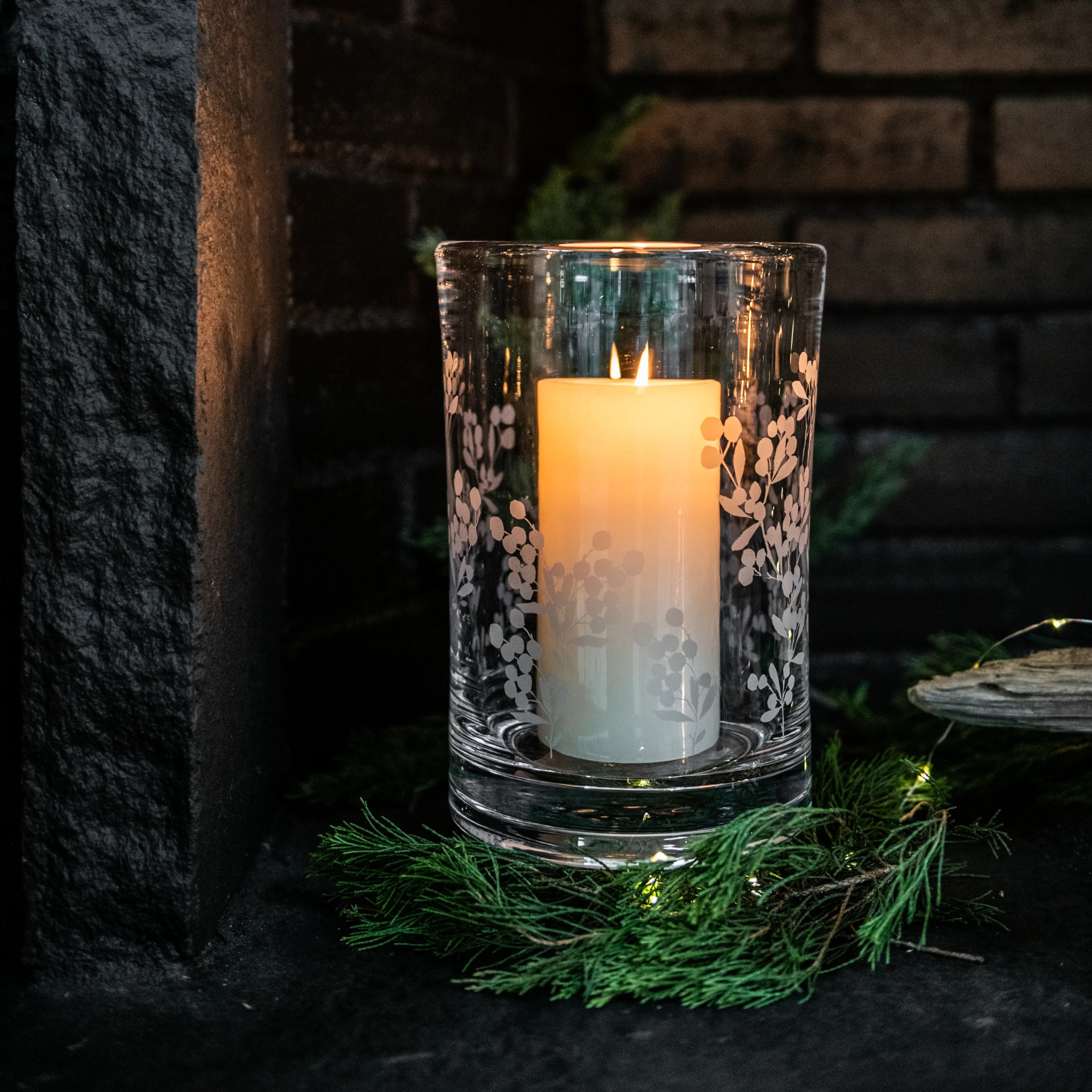 Clear glass hurricane with engraved berry and leaf pattern, containing lit cream-colored pillar candle.