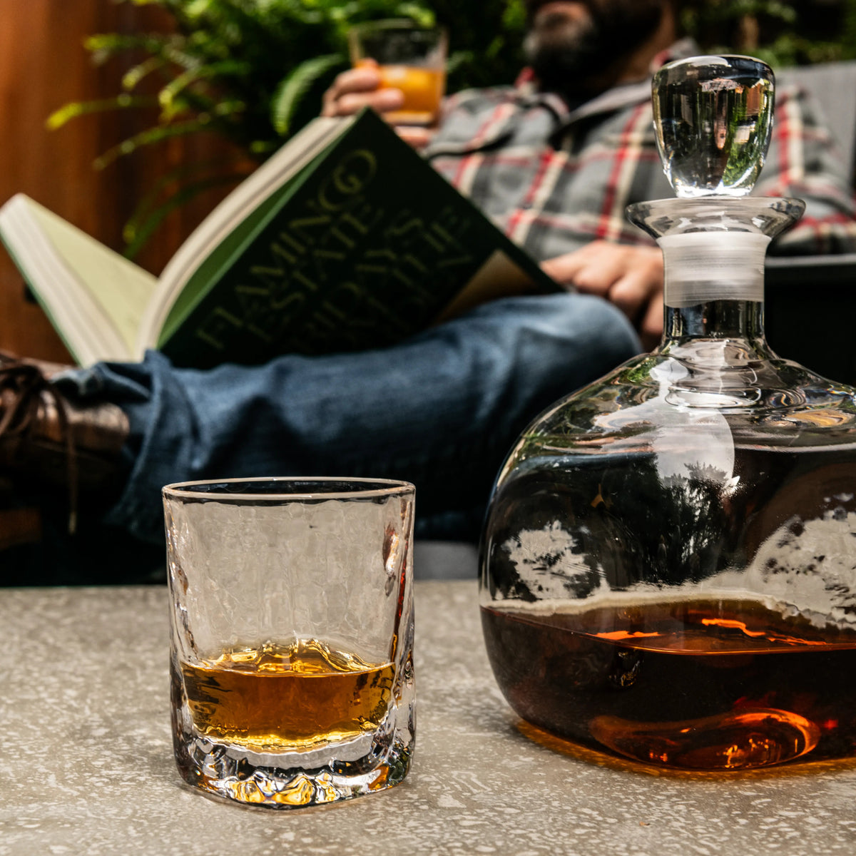 Clear double old-fashioned glass with amber liquid beside a stoppered decanter filled with brown spirit.