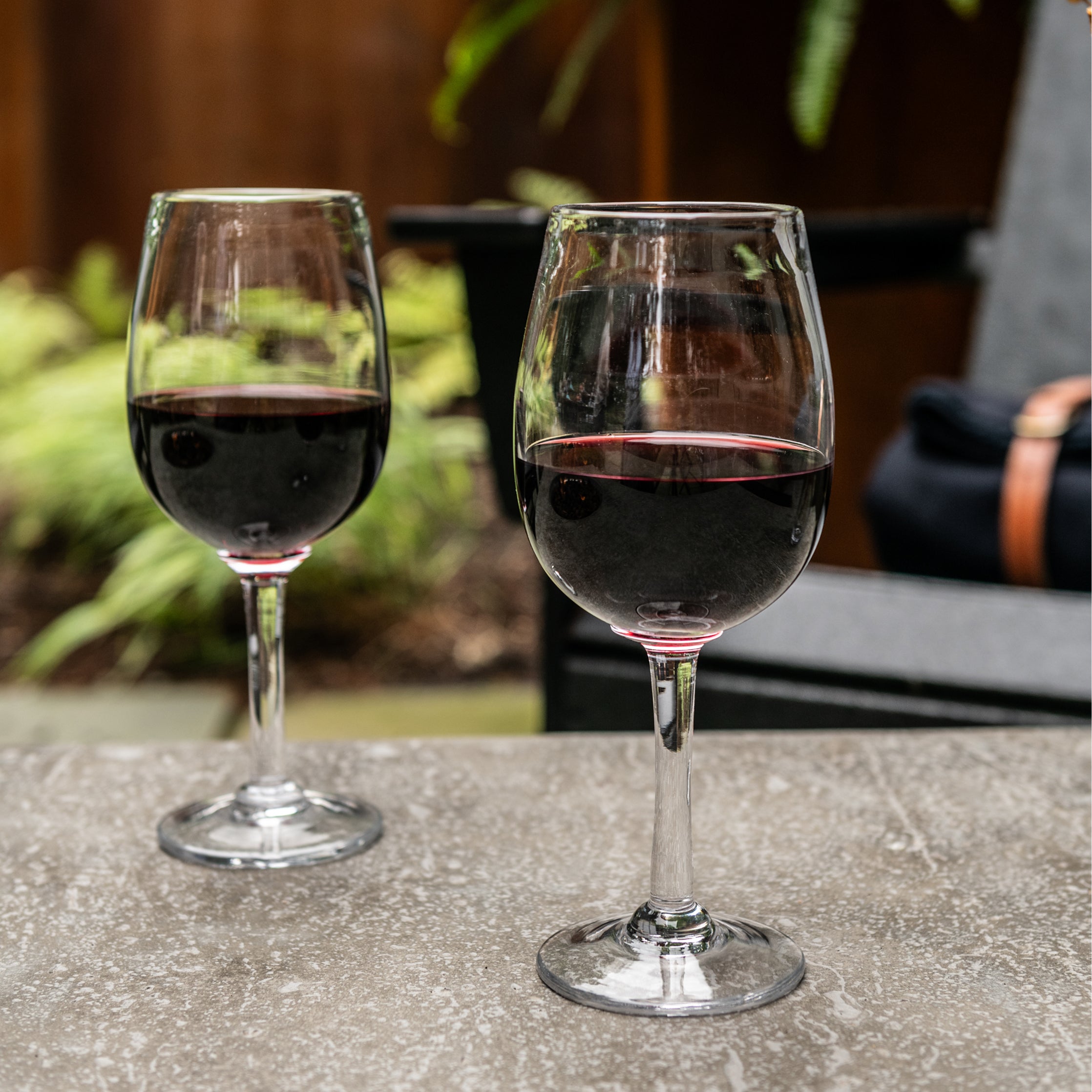 Two wine glasses filled with red wine on an outdoor surface with blurred greenery in background.