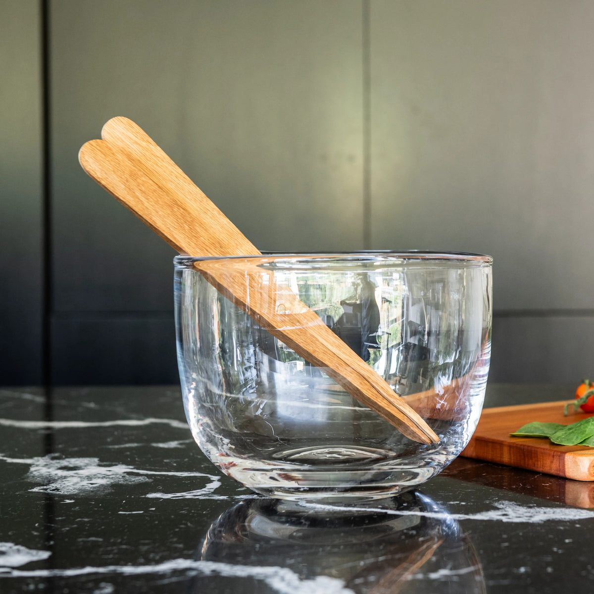 Two oak wood salad servers with tapered handles resting in a clear glass bowl on a dark marble surface.
