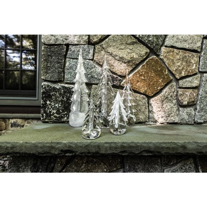 Three clear glass tree-shaped decorations with white frosted tops arranged on a stone ledge against a stone wall.