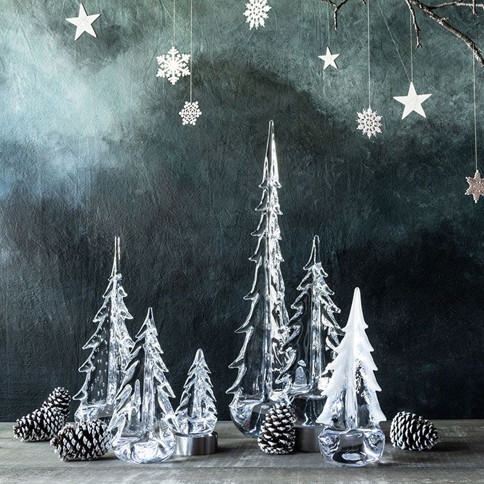 Clear glass evergreen tree figurines with pinecones and hanging snowflakes and stars on textured teal background.
