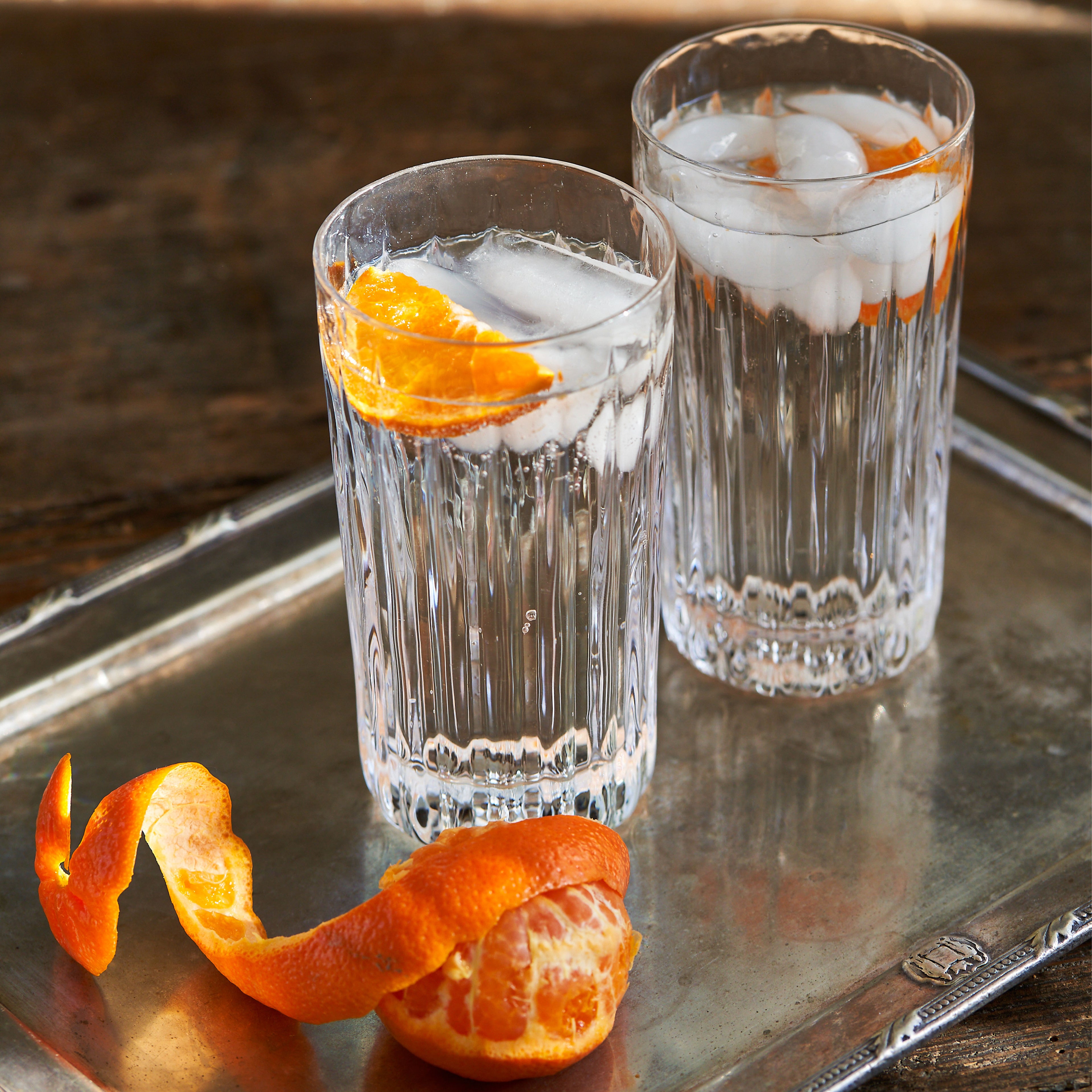 Two clear highball glasses filled with ice and clear liquid, garnished with orange slices, on a metal tray with fresh orange 