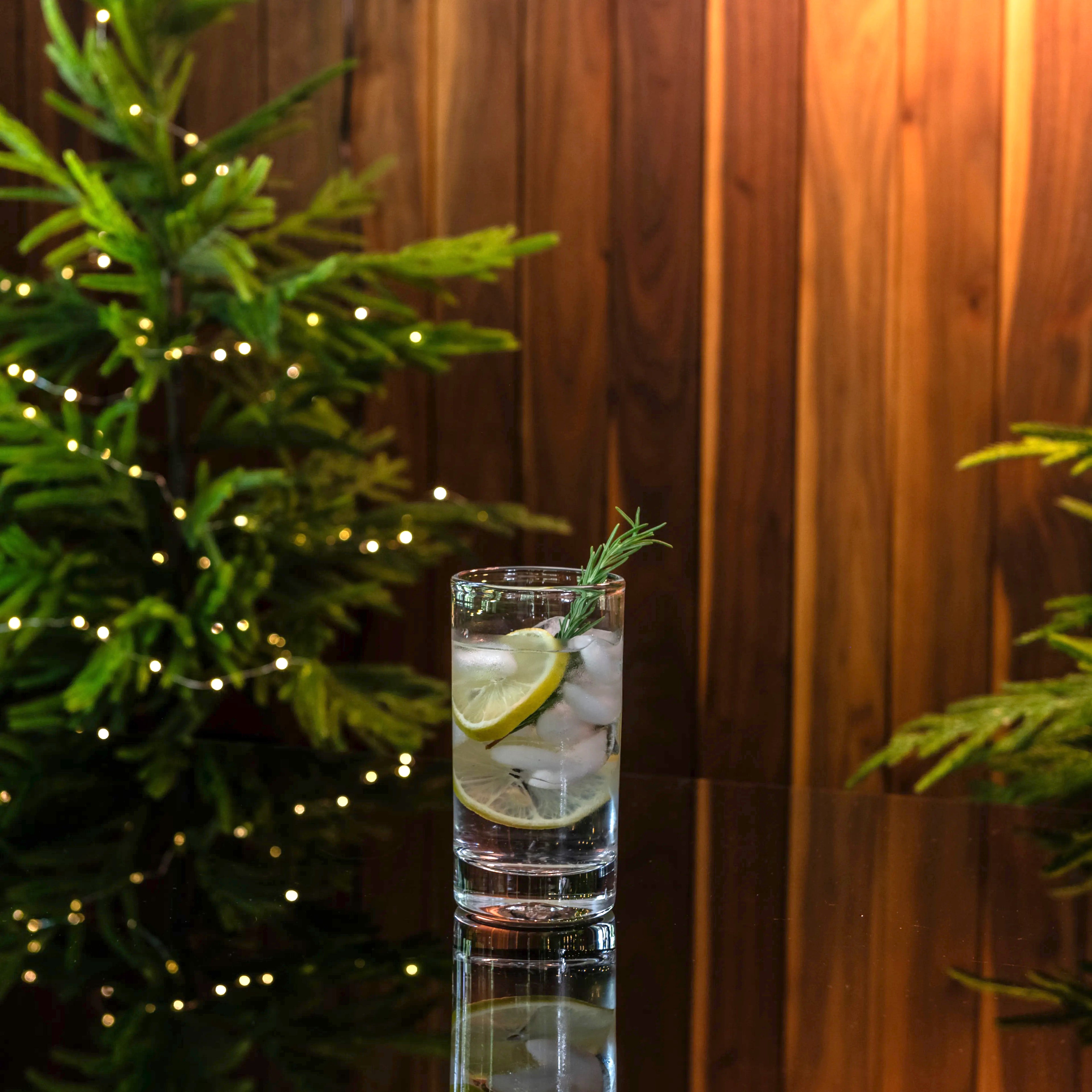 Clear glass tumbler filled with ice, lemon slice, and rosemary sprig on wood surface with greenery.