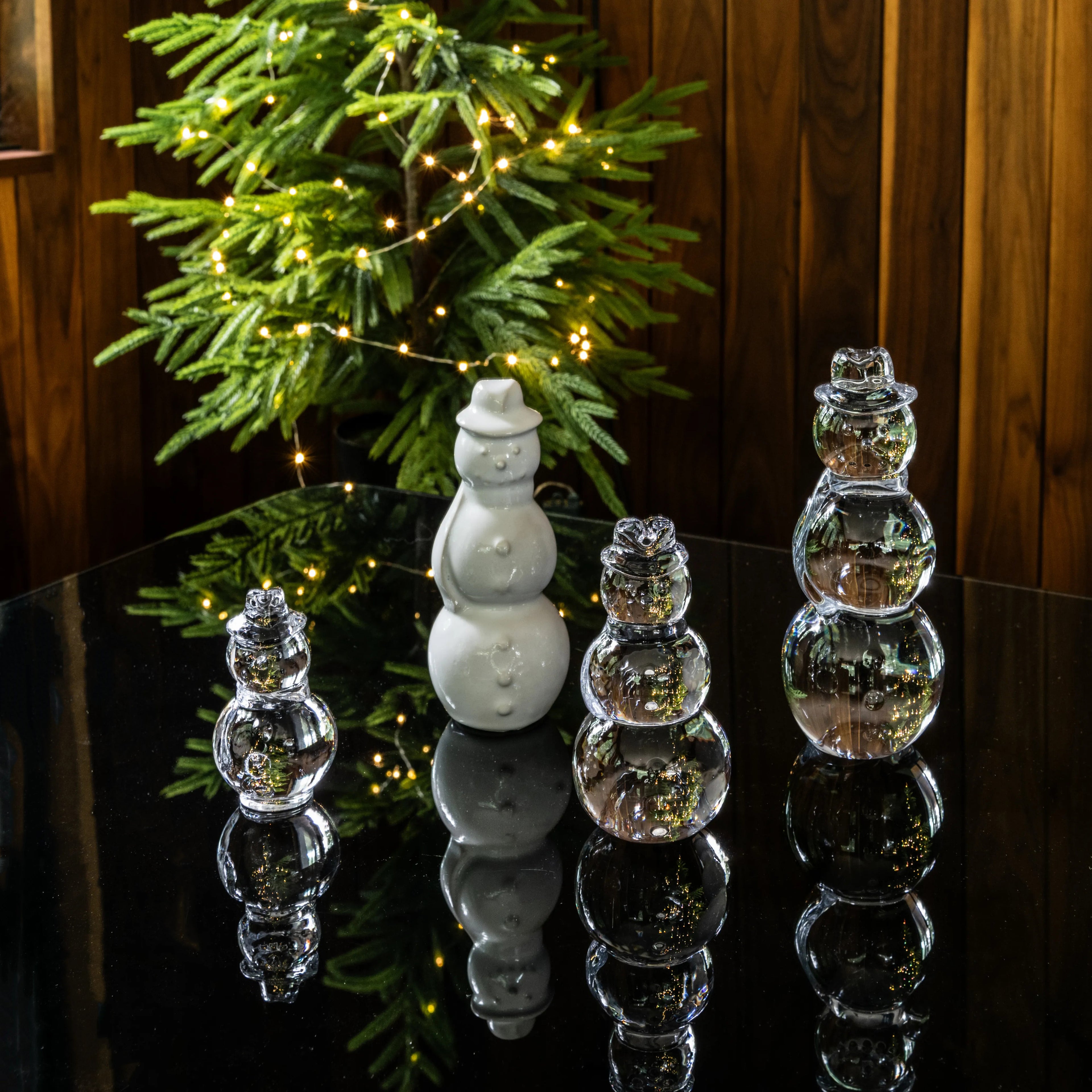 White ceramic snowman and clear glass snowmen figurines arranged on reflective surface with evergreen garland and lights.