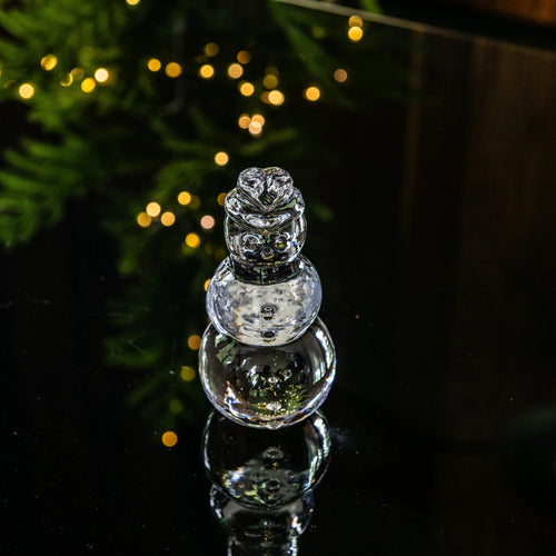 Clear glass snowman figurine with three stacked spheres and decorative top, displayed against green foliage and warm lights.