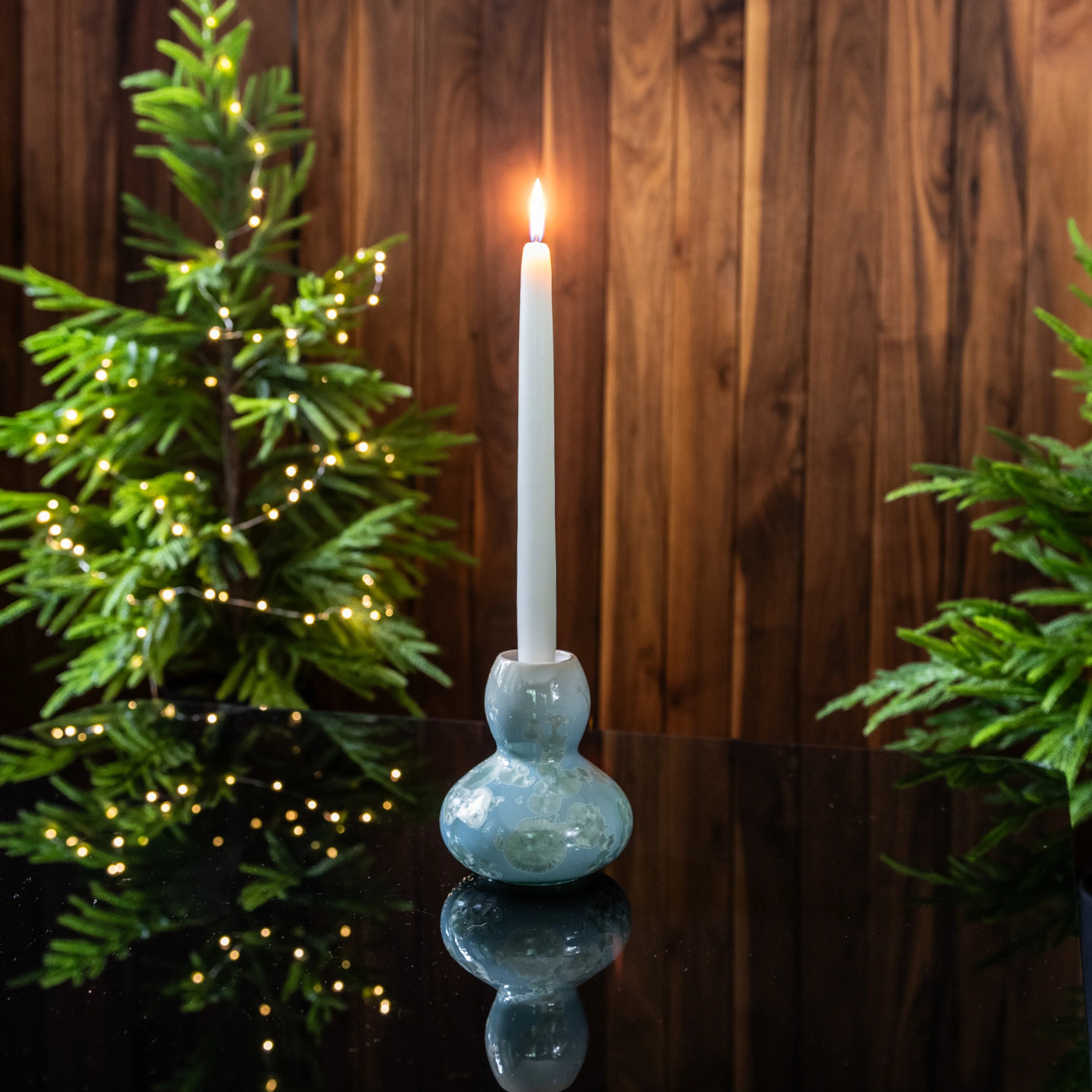 Blue pottery candlestick with bulbous base holding white lit candle, flanked by evergreen branches with small lights.