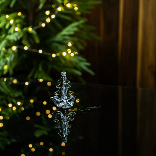 Clear glass tree-shaped ornament with gold accents, displayed in front of evergreen branches and warm white lights.