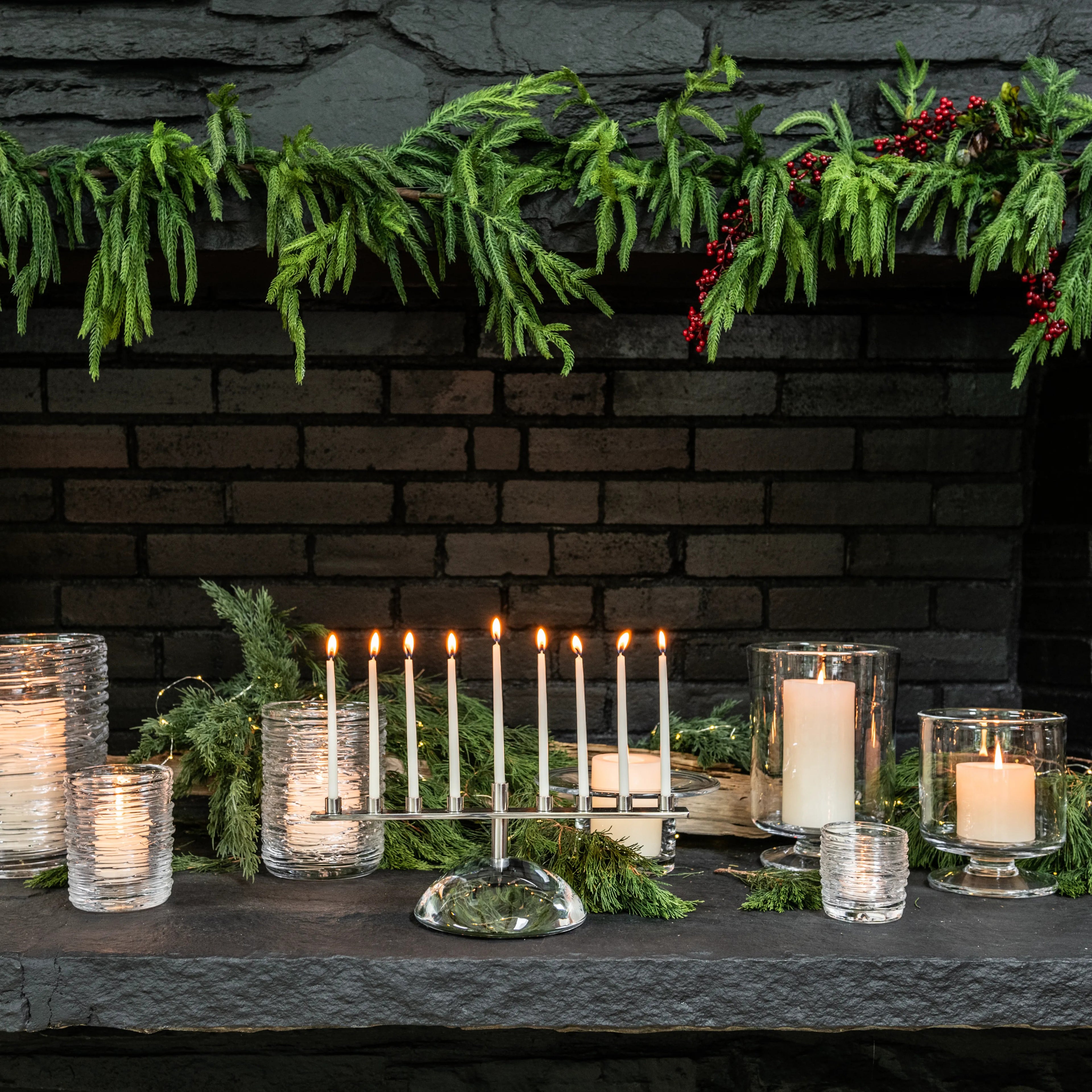 Silver menorah with nine lit candles displayed on dark fireplace mantel with evergreen garland and glass candleholders.