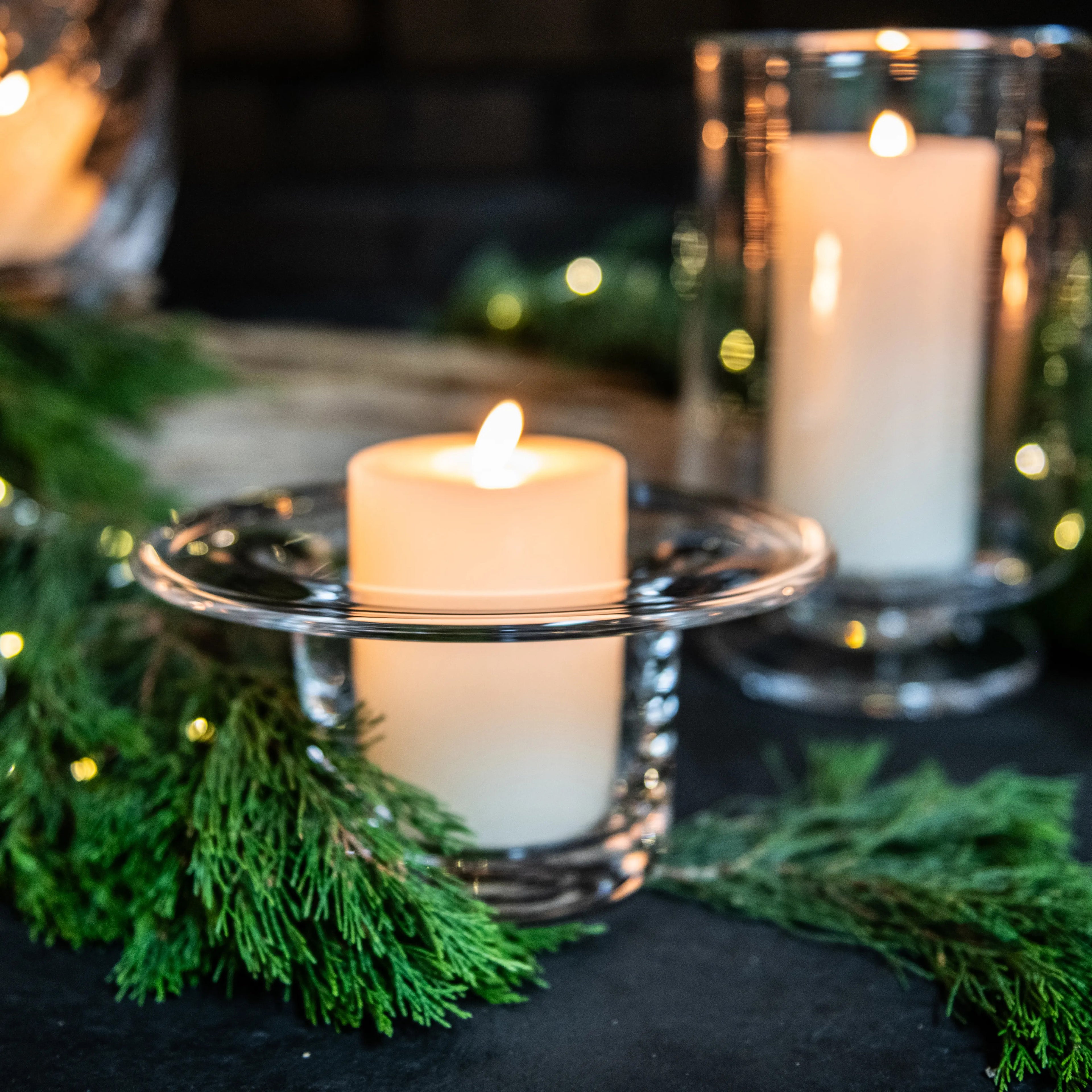 Lit tea light candles in clear glass holders surrounded by green evergreen branches and warm bokeh lights.