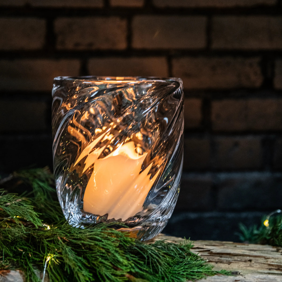 Clear textured glass candle holder with lit cream-colored candle surrounded by greenery against a brick background.