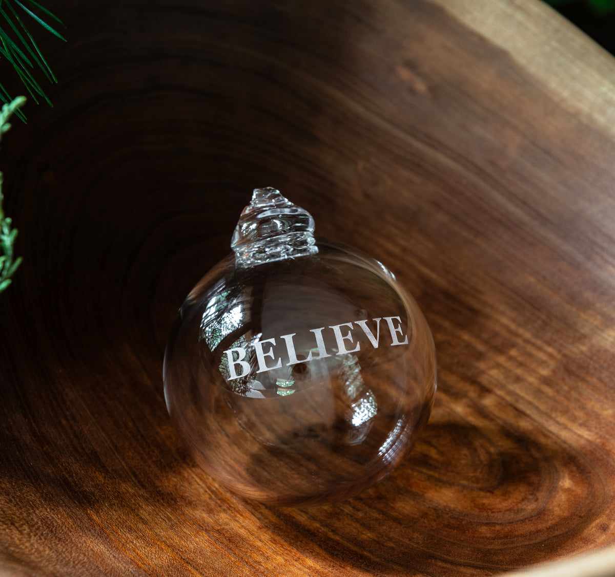 Clear glass round ornament with "BELIEVE" engraved text and metallic cap, displayed on wood surface.