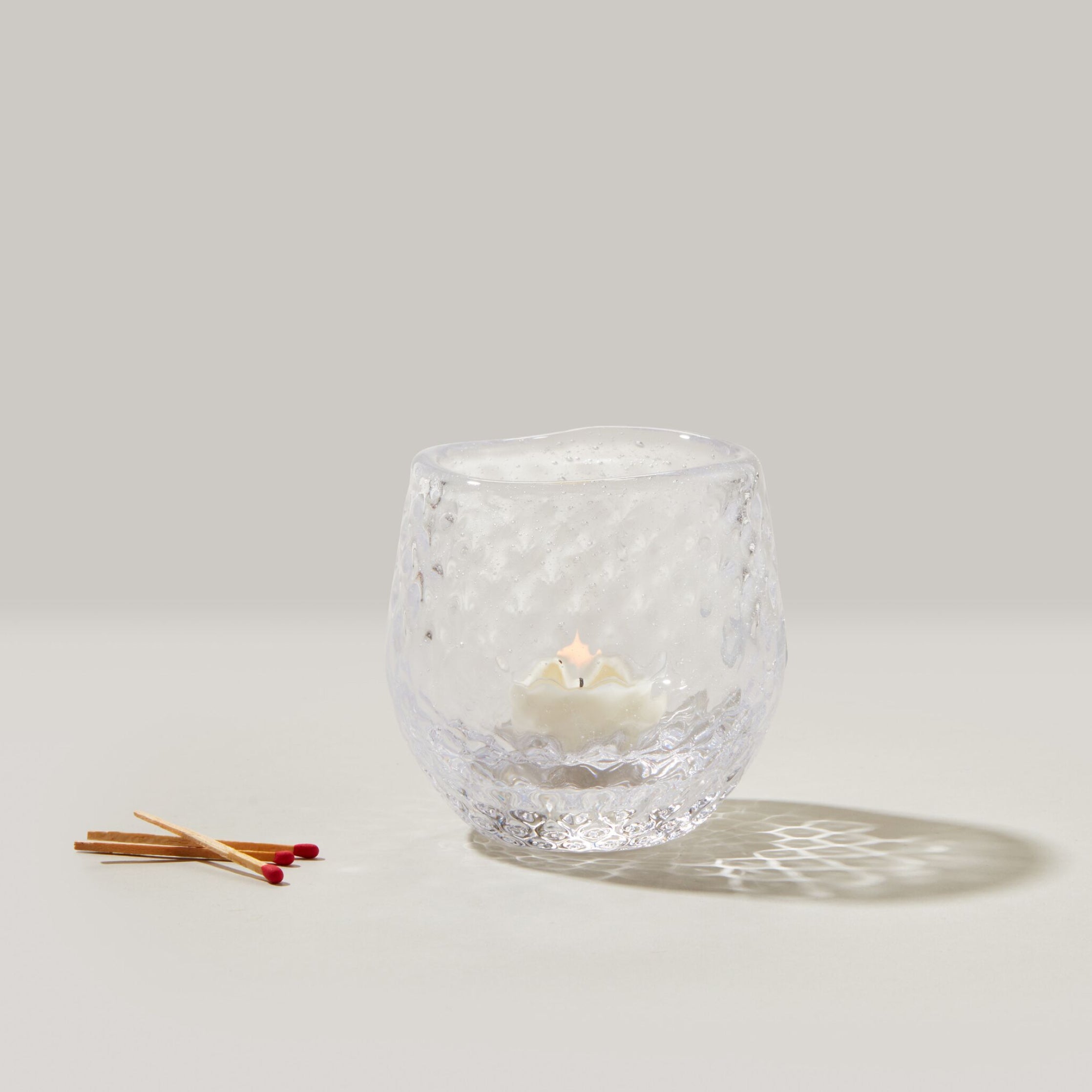 Clear textured glass tealight holder with lit cream candle inside and wooden matches beside it.