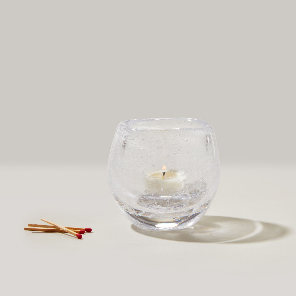 Clear glass tealight holder with lit white tealight candle inside and matches beside it.