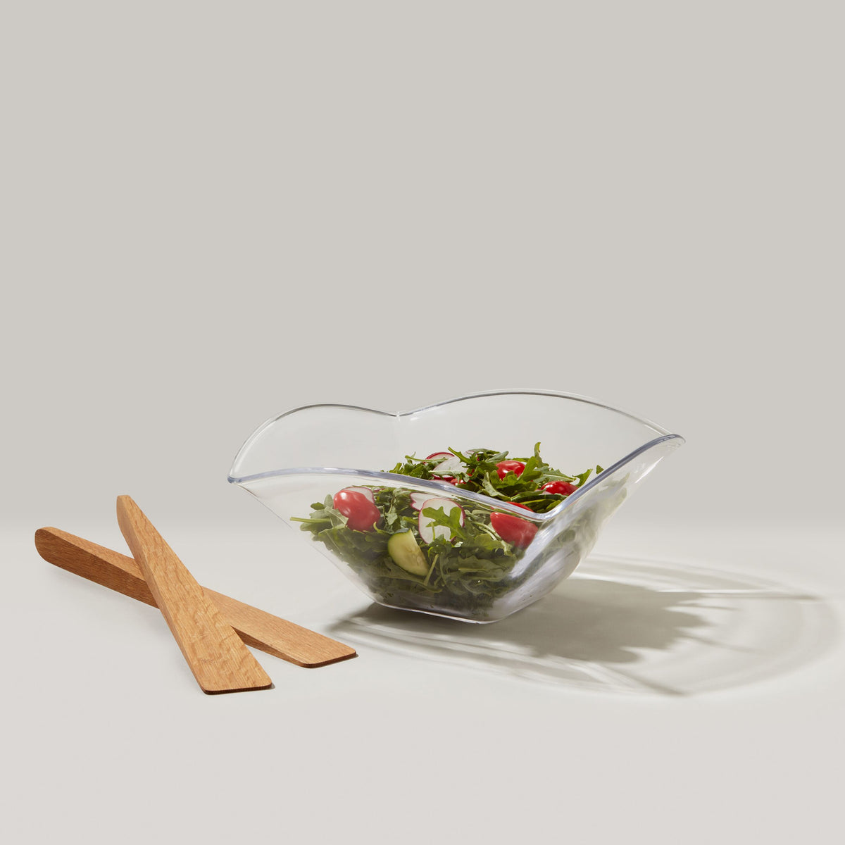 Clear glass serving bowl with wavy edges filled with mixed salad, accompanied by two wooden serving utensils.