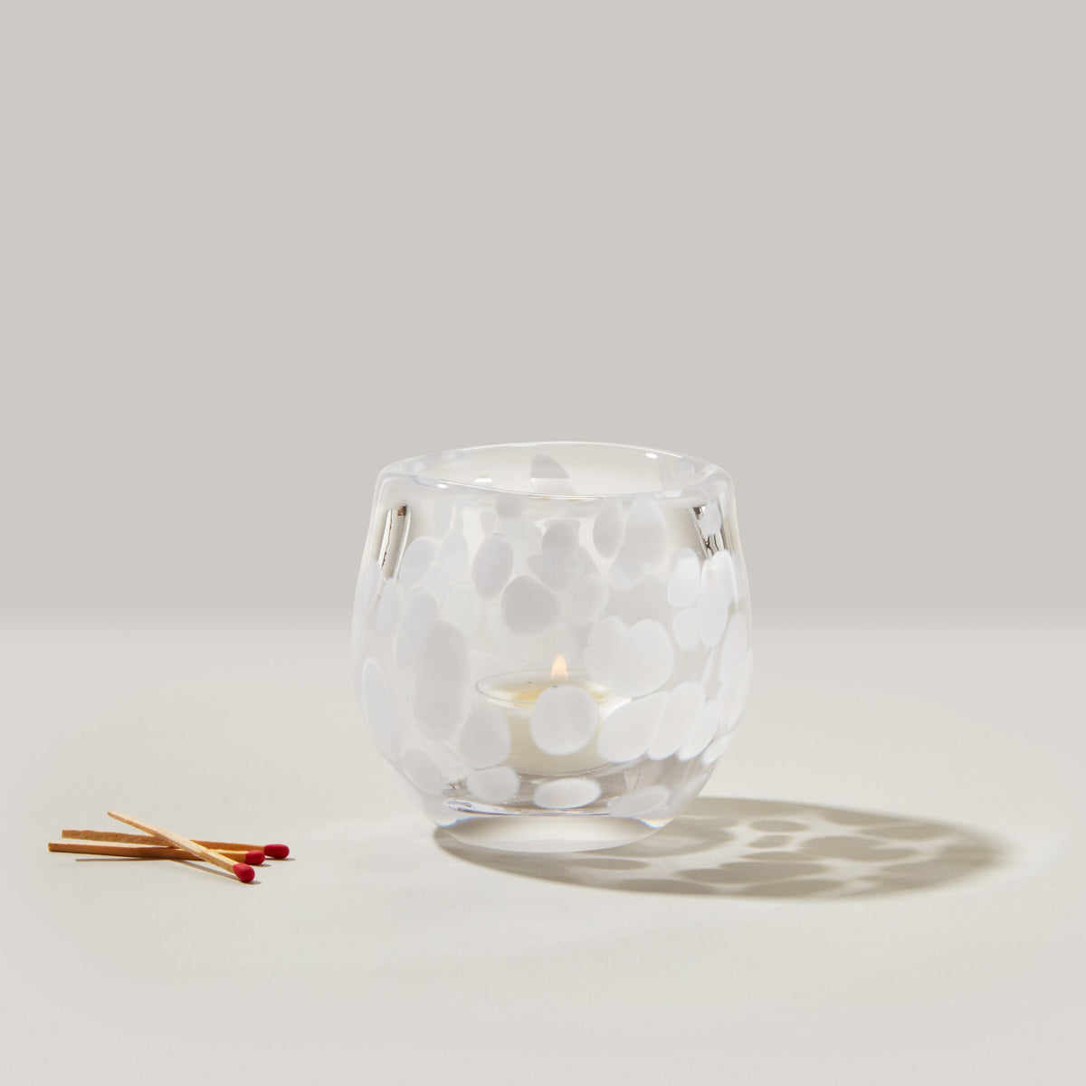 Clear glass tealight holder with white snowflake pattern and lit cream tealight inside, matches displayed next to it.
