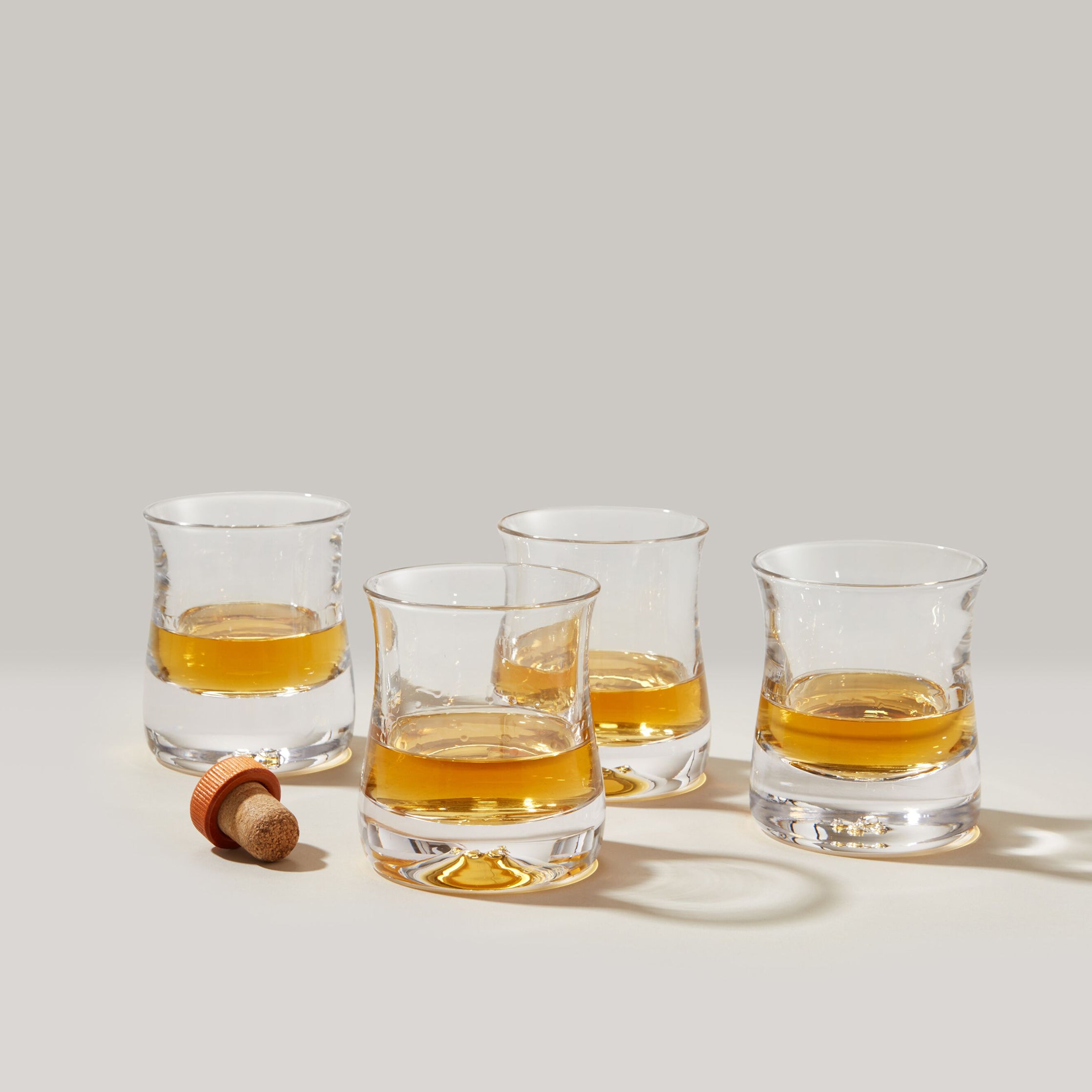 Four clear whiskey glasses filled with amber liquid and a cork stopper on a neutral background.