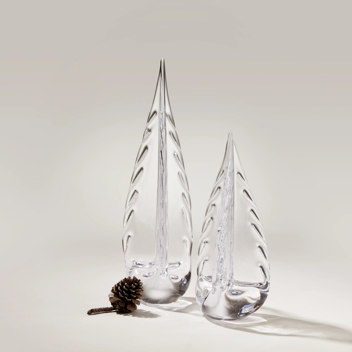Two clear glass evergreen tree figurines with ice-like texture, alongside a small pinecone on neutral background.