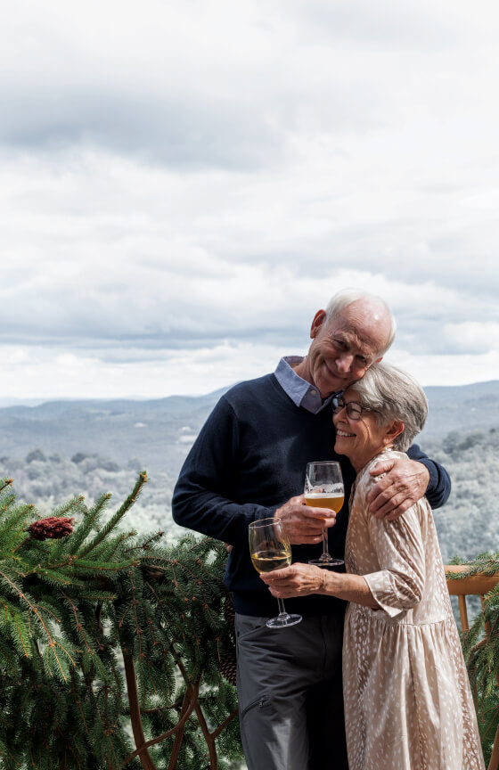 Elderly couple embracing with drinks, smiling, with a scenic backdrop.