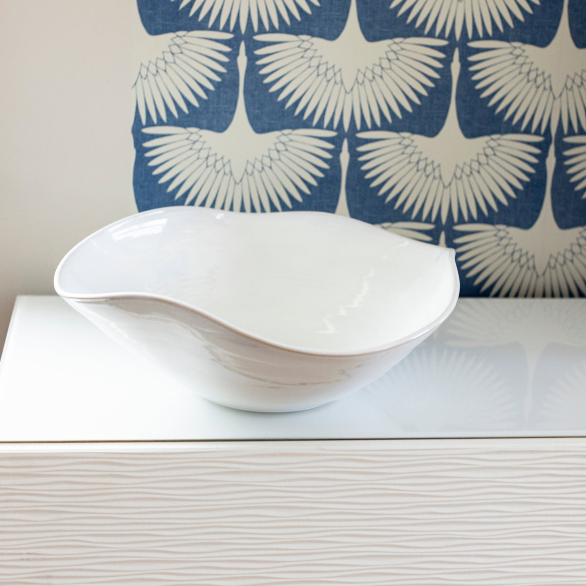 White ceramic bowl with curved organic shape displayed on white surface with blue patterned fabric background.