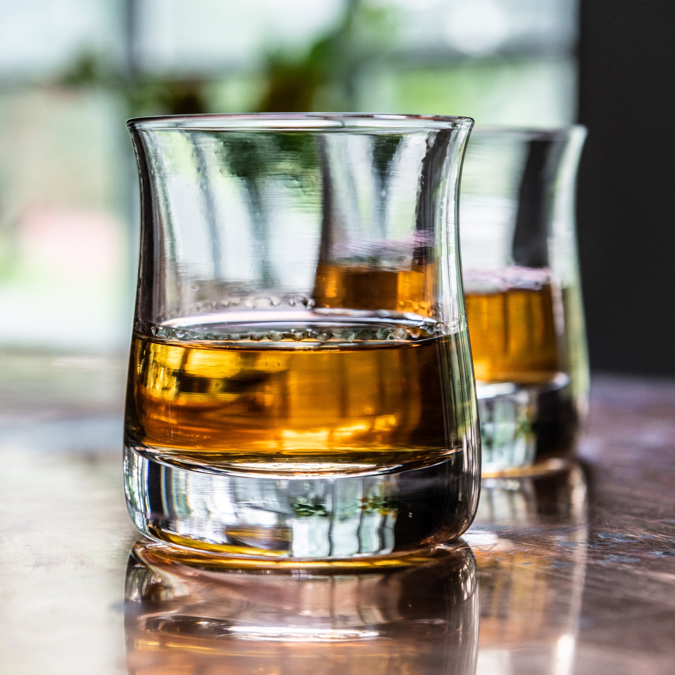 Two clear whiskey glasses filled with amber liquid and ice, displayed on a reflective surface with a blurred window backgroun