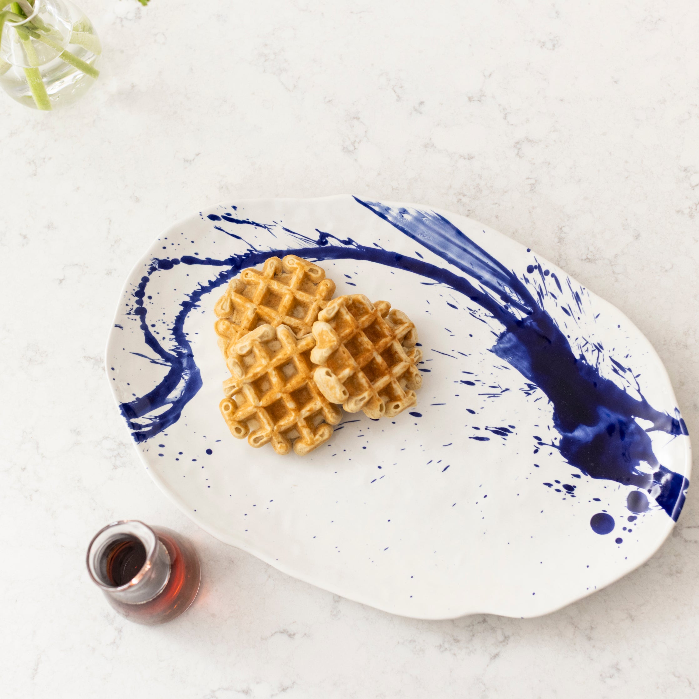 Oval white platter with blue brushstroke design, holding two waffles and a small glass of syrup.