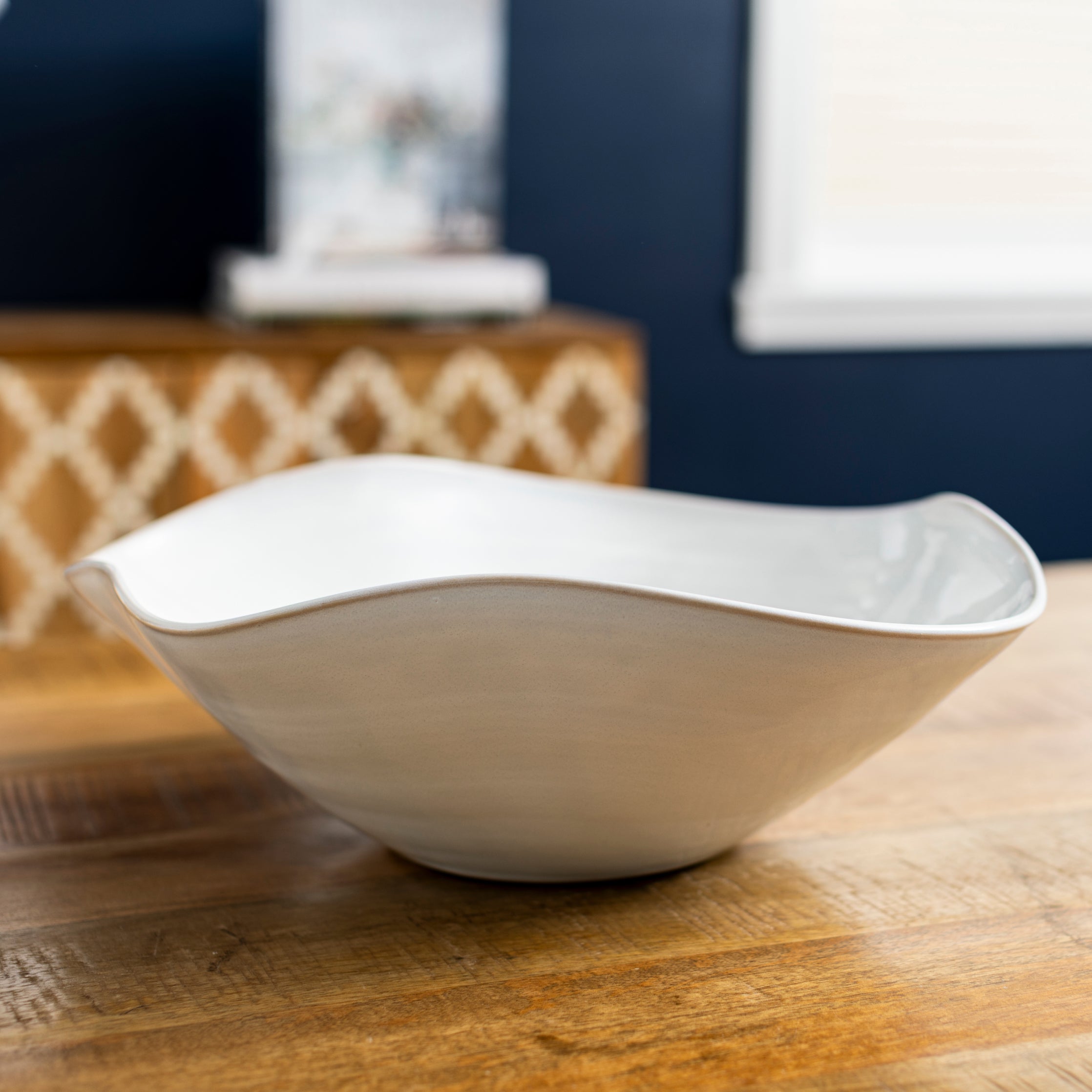 White ceramic bowl with curved wavy rim, displayed on wooden surface in home setting.