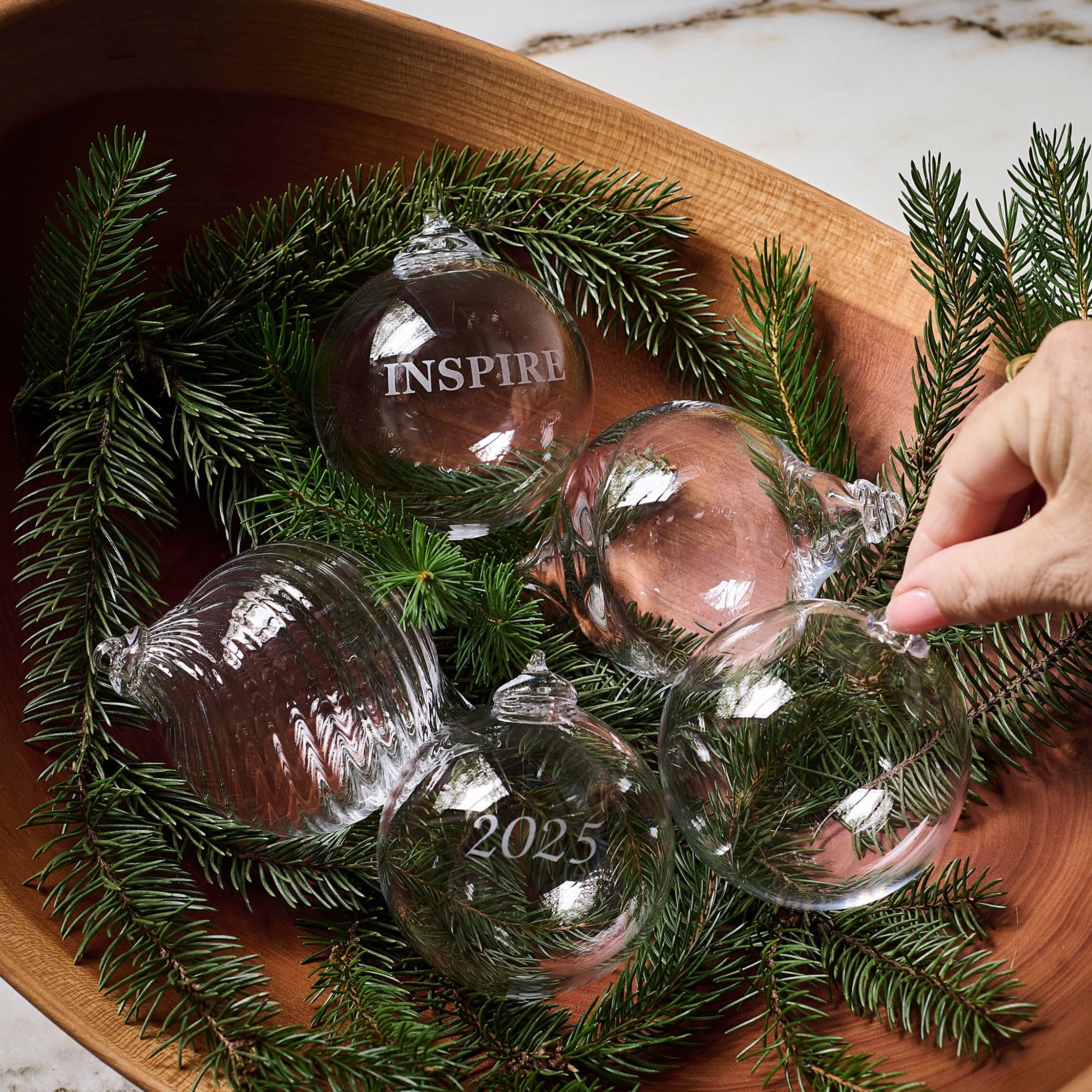 Round glass ornaments engraved with "Inspire" and "2025" nestled in evergreen branches in a wooden bowl.