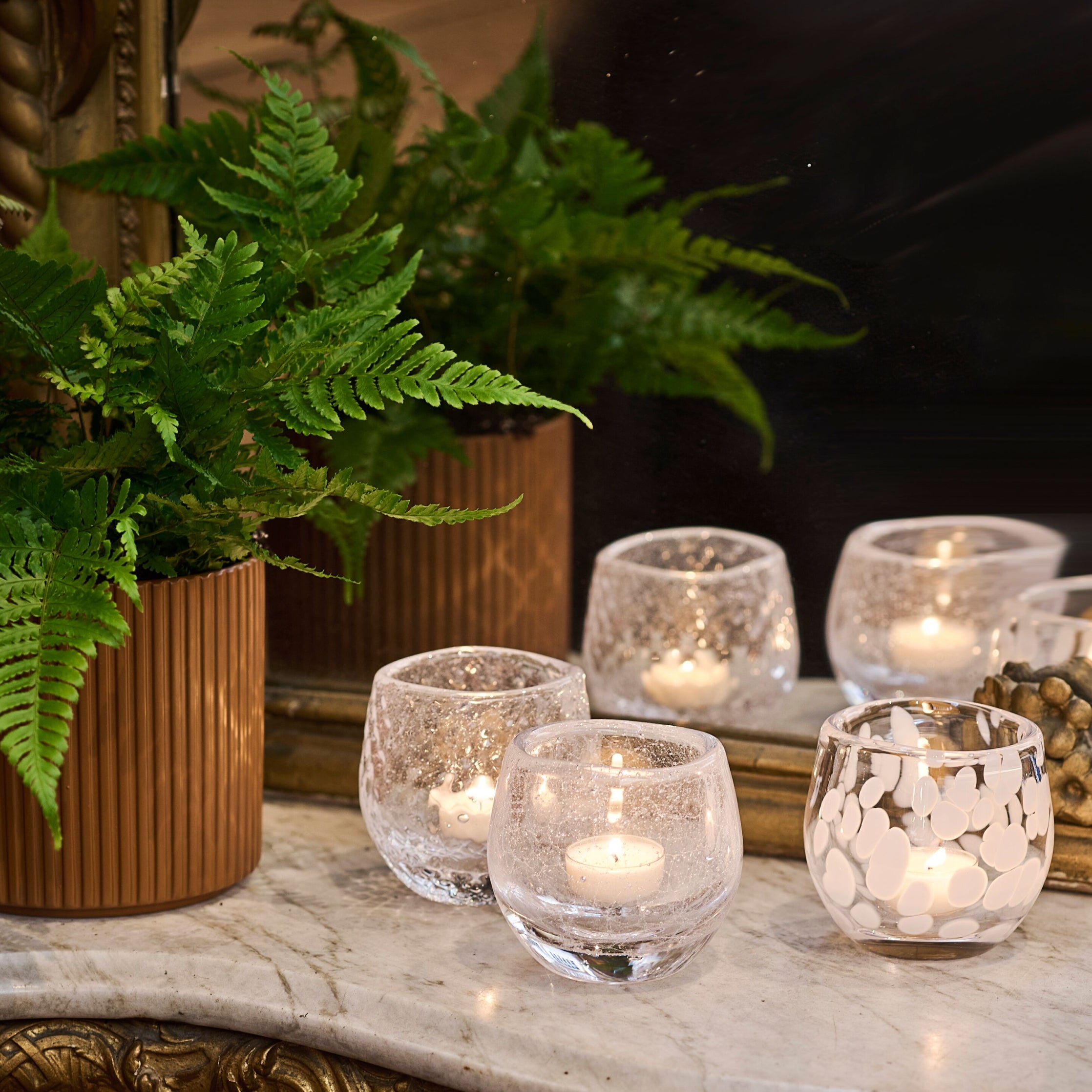 Clear glass tealight holders with lit candles, some featuring frosted or patterned surfaces, arranged on marble surface.