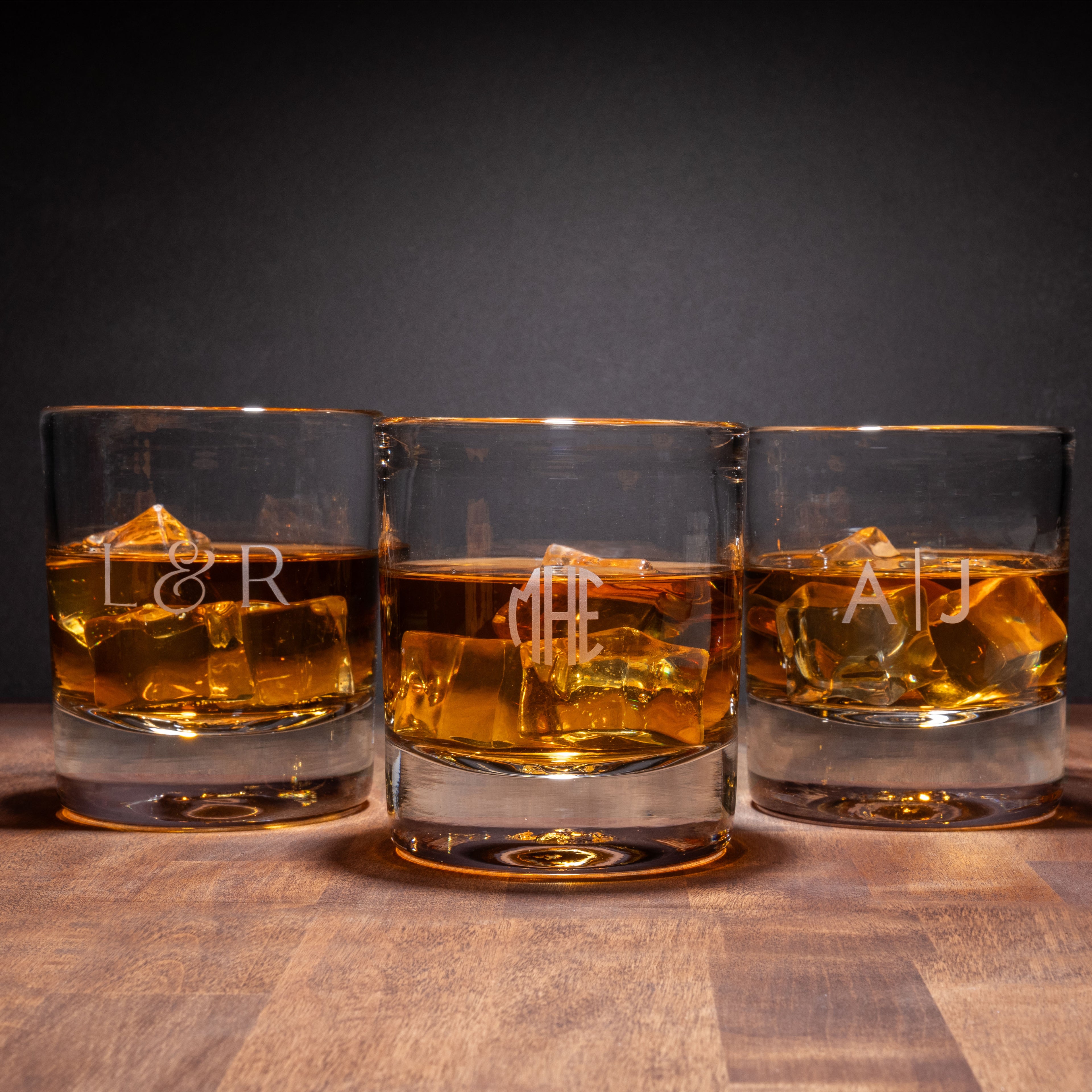 Three double old-fashioned glasses filled with amber liquid and ice cubes, each etched with initials.