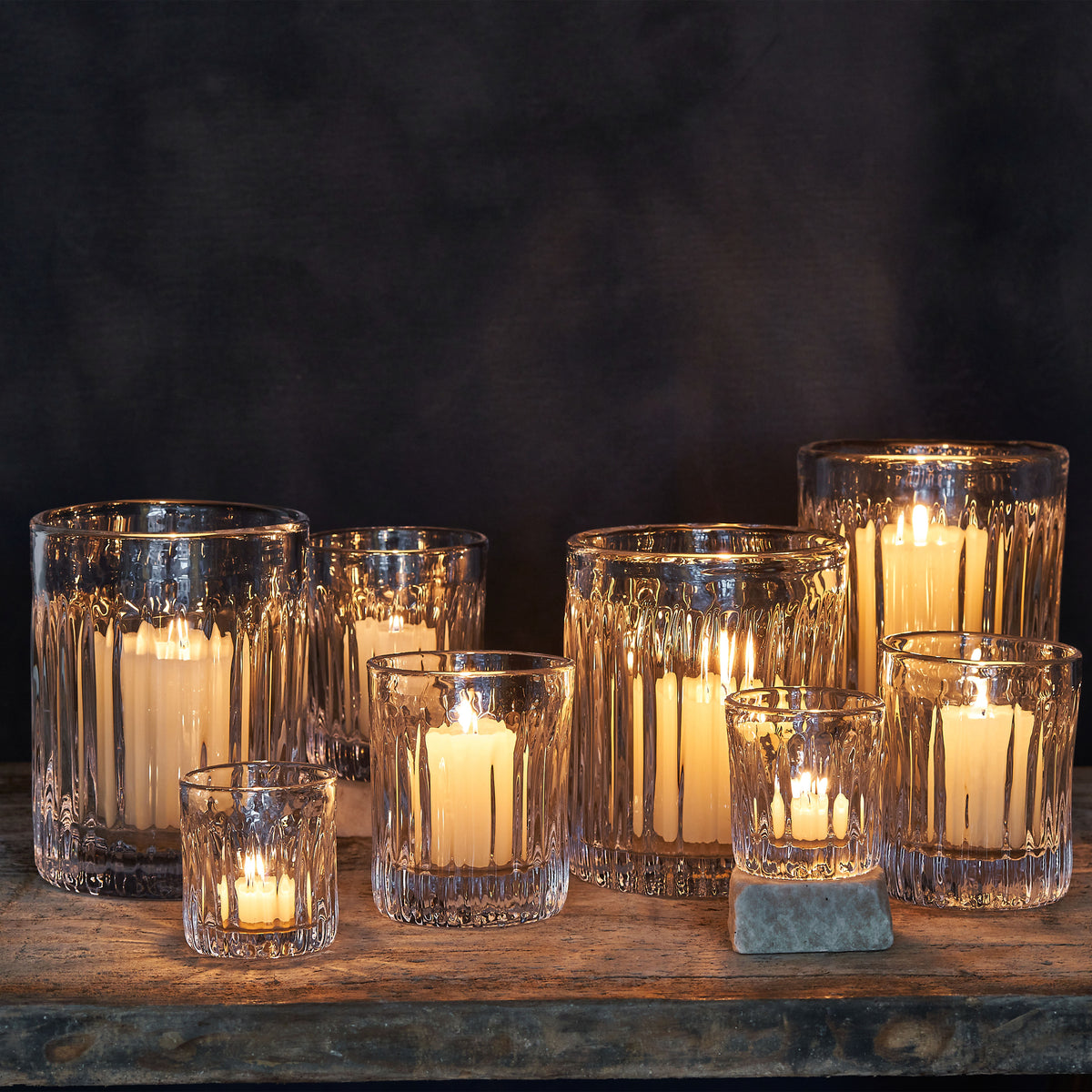 Seven clear glass hurricane candleholders with vertical ribbed texture containing lit cream-colored candles arranged on a woo